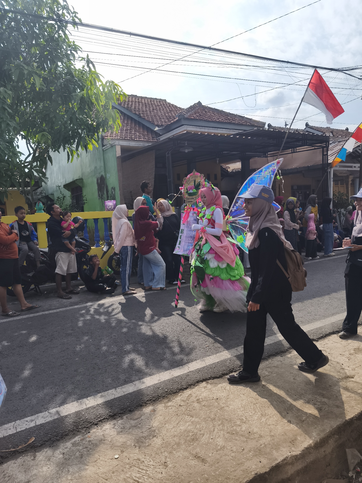 An outdoor daytime scene on a street in Kutoarjo, Indonesia, depicts a parade or procession. Several people are gathered on the side of the road, watching. In the center of the frame, a person in a colorful costume, resembling a fairy or butterfly, walks down the street. This individual is wearing a pink hijab and a voluminous dress in shades of pink, green, and white, with iridescent wings attached to their back. To the right of this costumed person, another individual wearing a black outfit and a light-colored cap walks in the same direction. Flags are visible in the background, including the Indonesian flag (red and white). The environment includes houses with tiled roofs and overhead power lines. The street is paved, and there's a concrete walkway on the right side of the frame. Some people are seated on motorcycles, observing the event.