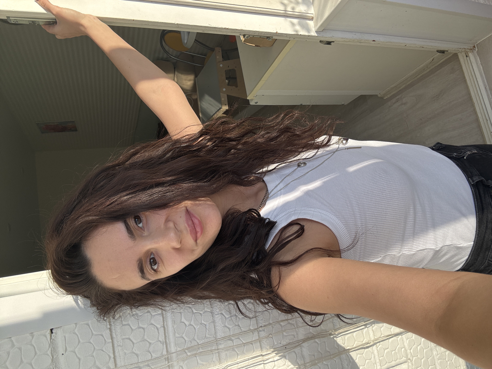 A young woman with brown hair and eyes is leaning out of a window. She wears a white ribbed tank top and black pants. Her left arm is raised, holding onto the top of the window frame. She is wearing a silver necklace. The window appears to be part of a building or structure with white trim. The scene takes place in daylight, with sunlight illuminating the subjects and environment. The location is Chaikovsky, Russia.