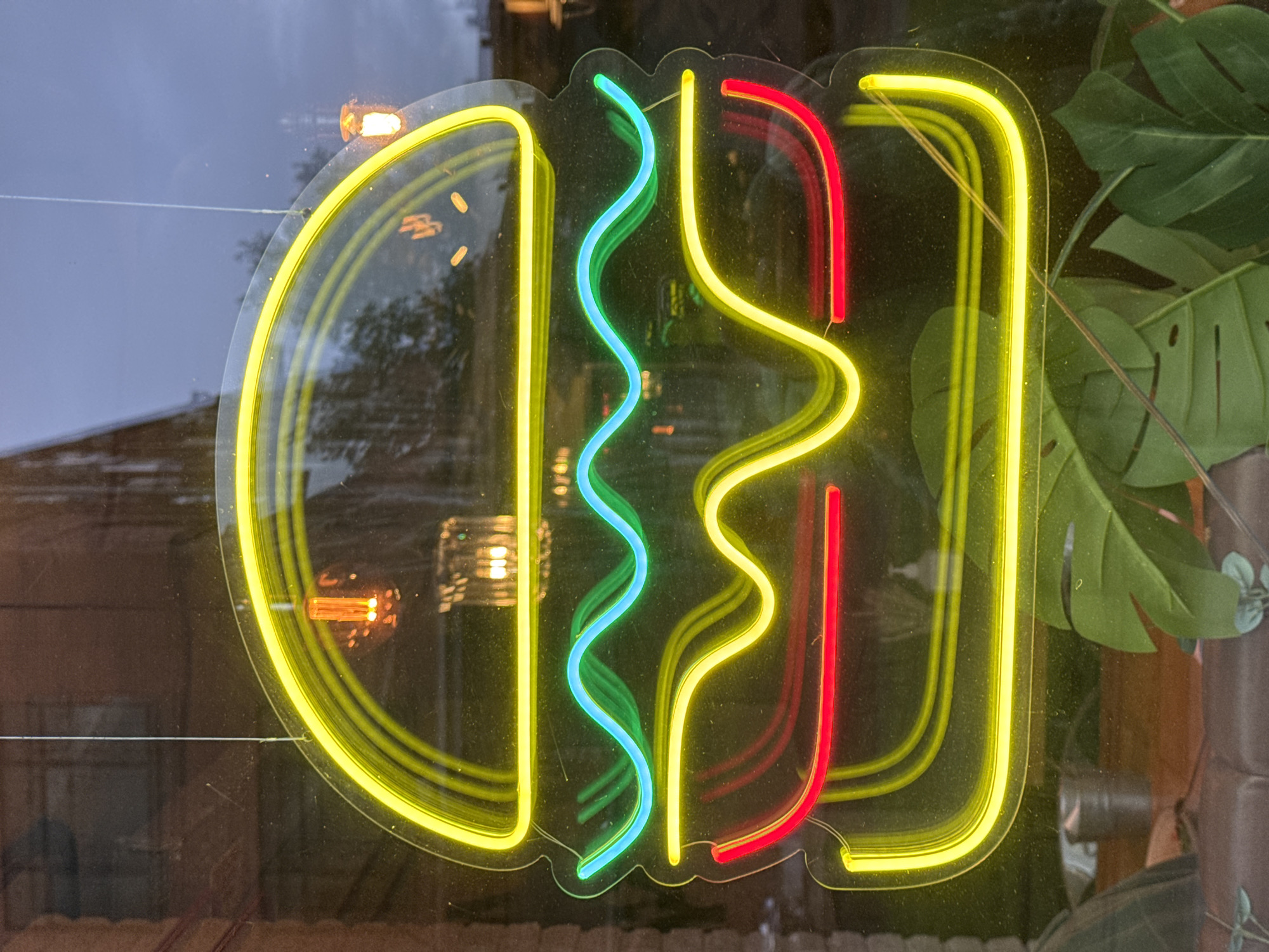 A close-up shot shows a neon sign of a burger in a window. The bun is represented by a double yellow line, the lettuce by a teal wavy line, the tomato by a red straight line, and the meat by a yellow wavy line. The window reflects a building across the street and is placed next to a green plant.