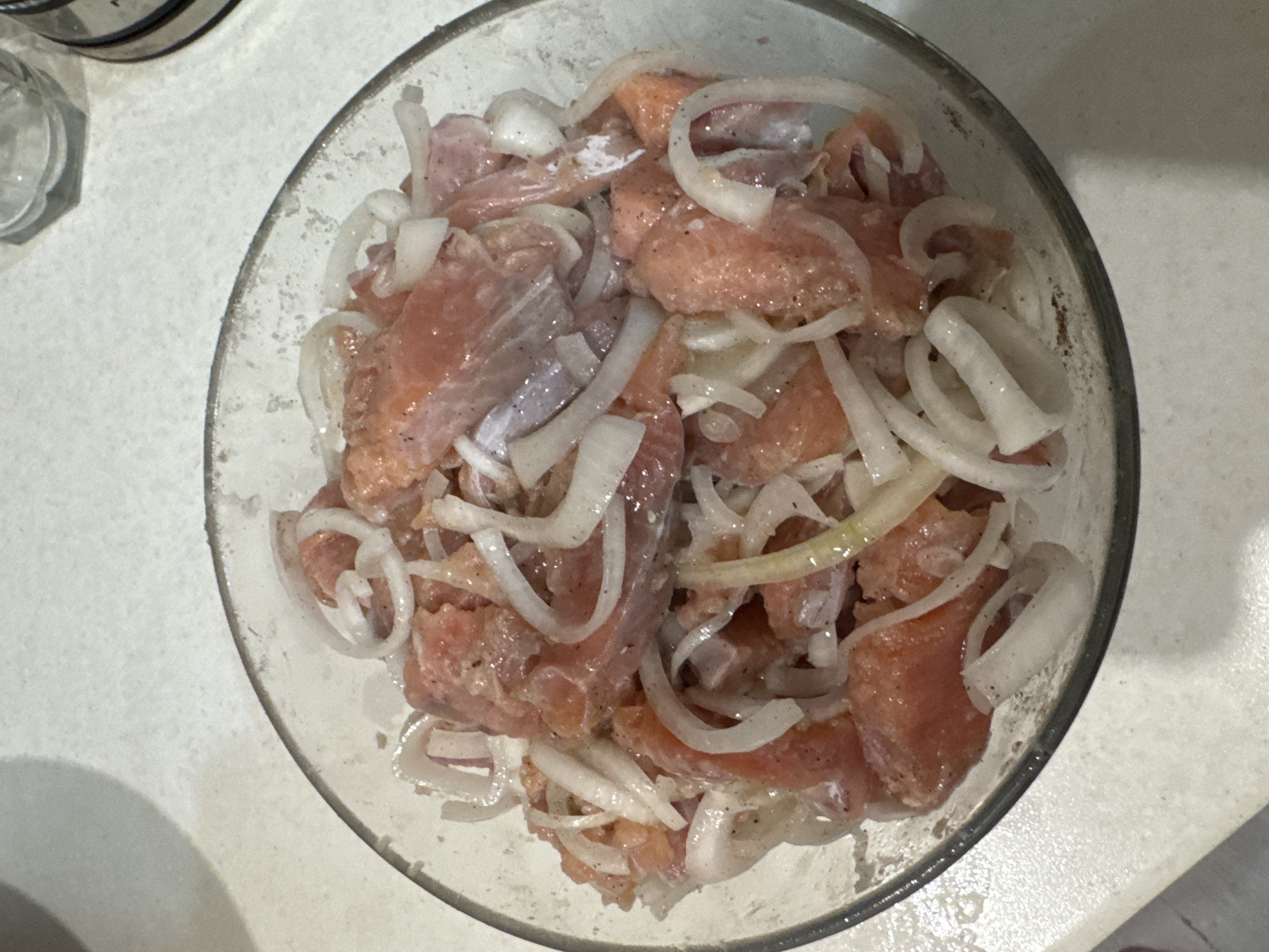 A bowl filled with sliced, raw fish (possibly salmon) and thinly sliced onion rings sits on a light-colored countertop. There is a jar or container to the upper left of the bowl. The scene appears to be indoors. The location cannot be determined from the image.