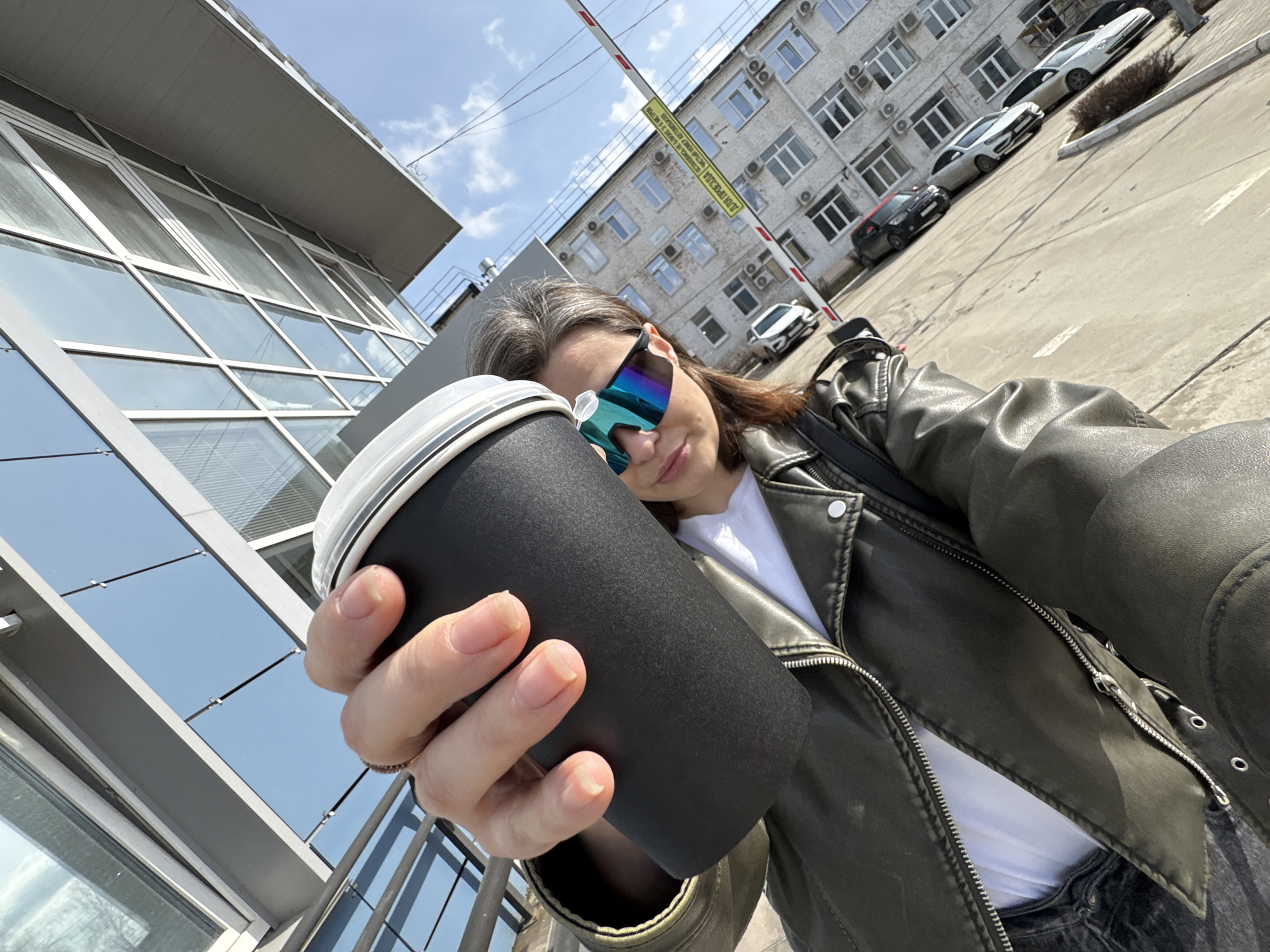 A woman holds a black coffee cup in a selfie in what appears to be an urban setting in Chaikovsky, Russia. The location is suggested by the yellow sign in the background, which features Russian text.
The woman wears reflective sunglasses, a white t-shirt, and a leather jacket. Her hair is partially gray. She is holding the coffee cup out in front of her.
Behind the woman are multiple cars parked in front of a light-colored brick building with many windows. Air conditioning units are visible on the building's exterior. A gray building with glass windows is visible to her left. Overhead is a metal barrier. The sky is partially cloudy.