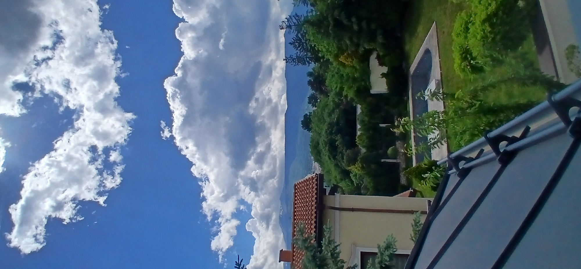 The image shows a view from what appears to be a residential area. In the foreground, there is a partial view of what could be a patio or awning structure, composed of light-colored panels and dark supporting beams. Behind this, the scene opens up to reveal a house with a tan-colored facade and a red tile roof. Beyond the house is a densely vegetated area with various trees and shrubs, suggesting a garden or parkland. In the distance, there are mountains, partially obscured by clouds. The sky takes up a significant portion of the image and is filled with billowing white clouds against a blue backdrop.
There are no people visible in the scene. The image focuses on the natural and built environment. The details suggest a calm and possibly affluent residential area.
Given the presence of mountains and the architecture, it's plausible this location could be in a suburb of Sofia, Bulgaria. The presence of certain types of foliage would confirm this, but the current image does not provide a clear, detailed view of them.