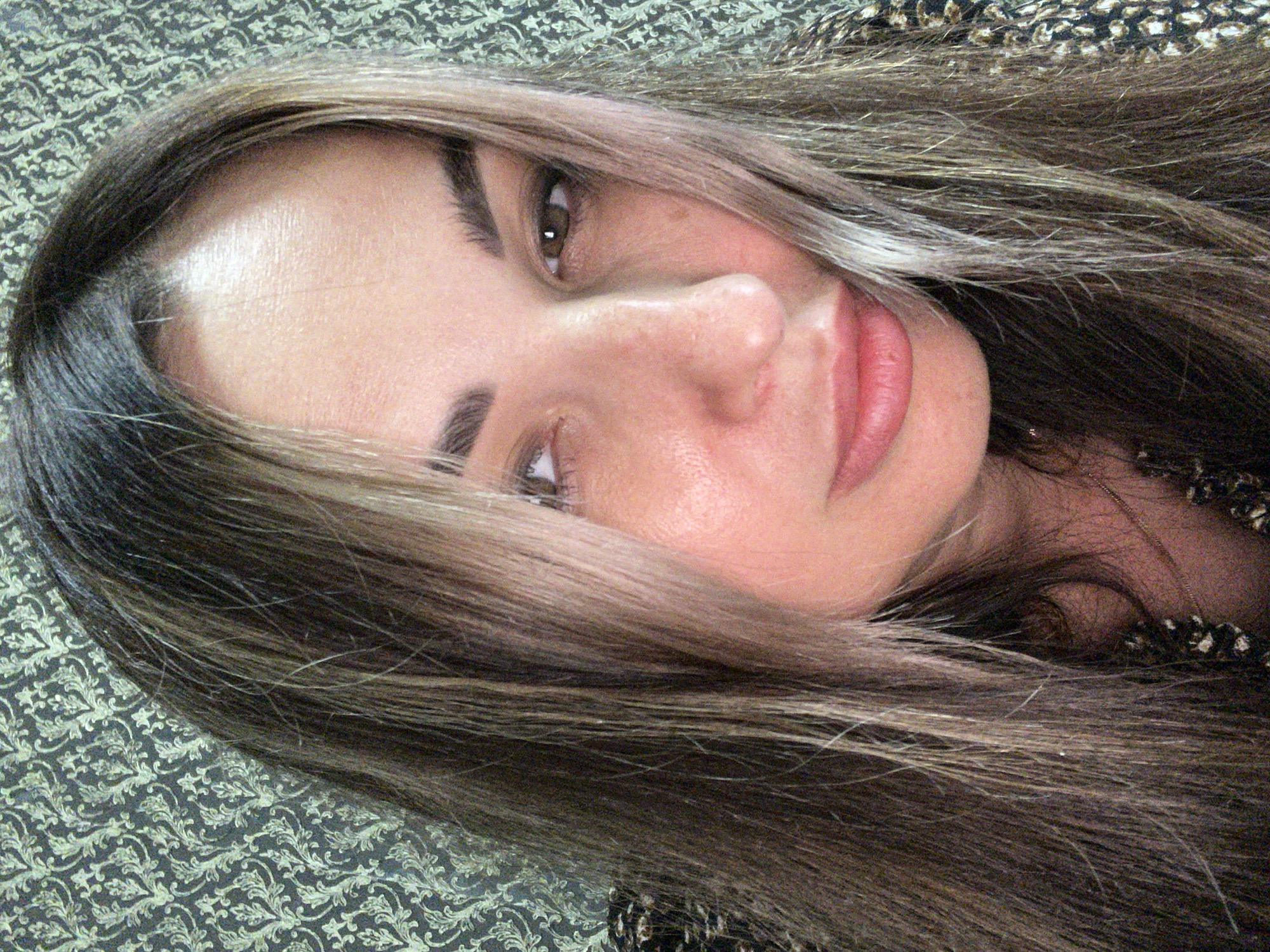 A medium close-up shot captures the face of a woman with long, straight, light brown and blonde hair that partially obscures her face, a textured patterned wallpaper in the background, and her upper chest and shoulder wearing a black and gold patterned top with a delicate gold necklace. The woman's face is turned slightly to her left, with her eyes looking to her left, her nose pointed forward, and her mouth slightly open. Her skin appears smooth with a faint blush on her cheeks and lips. She has dark eyebrows and visible strands of hair in front of her forehead and eyes. The lighting casts a soft glow on her face. The overall scene is a self-portrait captured indoors.