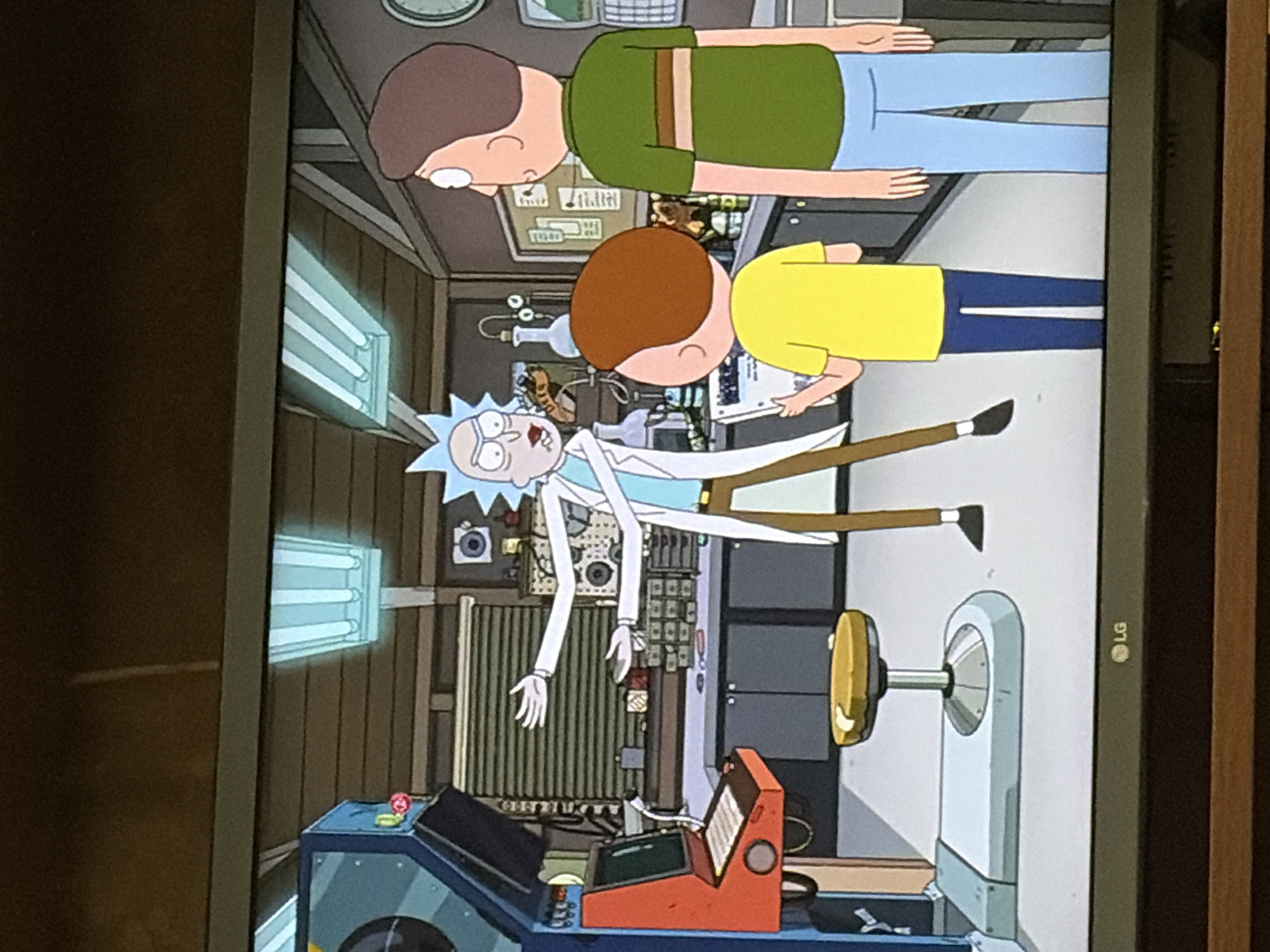 The image depicts an indoor scene, possibly a garage or workshop, where three animated characters are present. On the left, a character resembling Rick Sanchez from the show "Rick and Morty" is seen gesturing with his hands, wearing a lab coat over a white shirt and brown pants. In the center, a younger character, likely Morty Smith, is wearing a yellow t-shirt and blue pants, holding a white folder. To the right, an adult male character with brown hair and wearing a green t-shirt with horizontal stripes and blue pants stands looking forward. In the background, there are workbenches, cabinets, scientific equipment, and what appears to be an arcade-style machine. The lighting suggests fluorescent overhead lights. The overall environment appears functional and somewhat cluttered, consistent with a laboratory or workshop setting. There are no clear indicators of a specific city or outdoor environment.