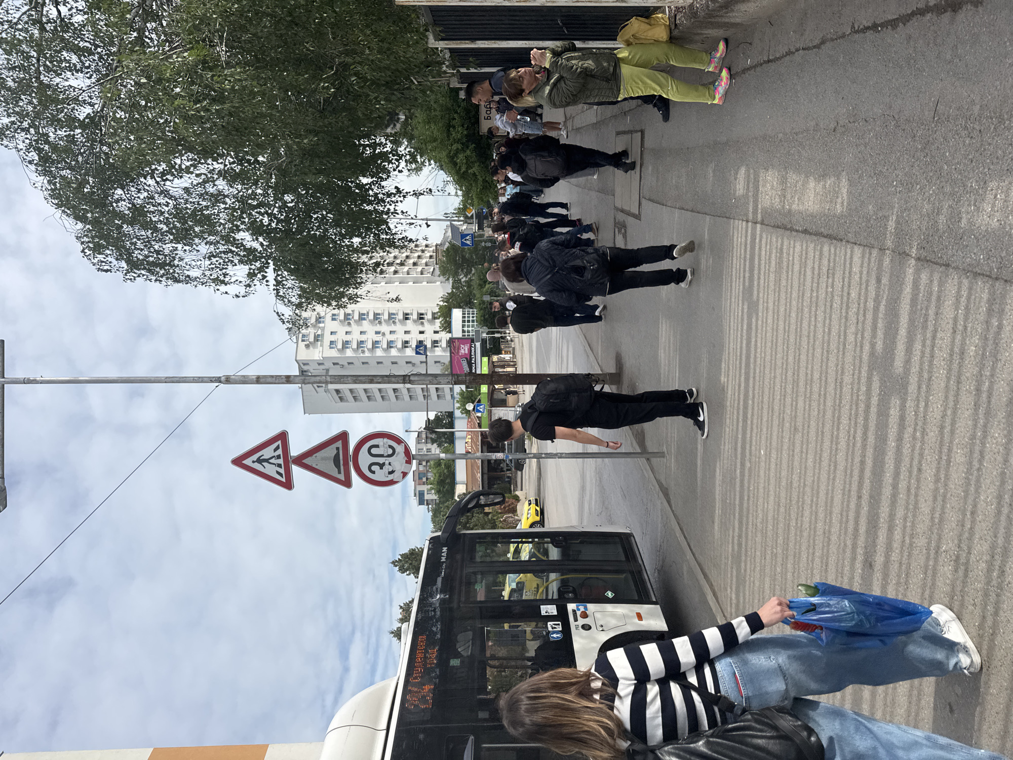 The image shows a street scene in Sofia, Bulgaria. A line of people are waiting on a sidewalk next to a city bus. The bus is white with black trim and "MAN" is written on the side. A woman is sitting in the bus. There are pedestrian crossing and speed limit (30 km/h) signs next to the bus stop. Beyond the bus stop, there is a yellow taxi cab parked on the street. In the background is a line of buildings. A large tree is along the sidewalk. The sky is partially cloudy.