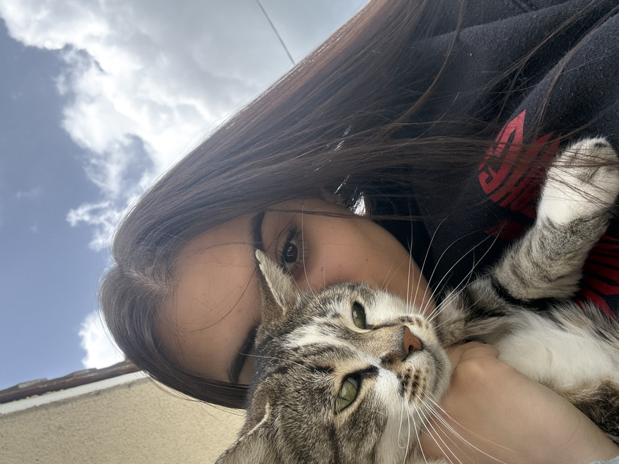 A medium close-up portrait shows a young woman holding a tabby cat. The woman has long, dark brown hair, and her face is partially visible. The cat is grey and white with green eyes. The woman wears a black hoodie, with what appears to be a red logo or design on the shoulder. The background includes a cloudy blue sky, and a beige wall can be seen at the bottom. There's no indication of location in the city of Polena, Bulgaria.
