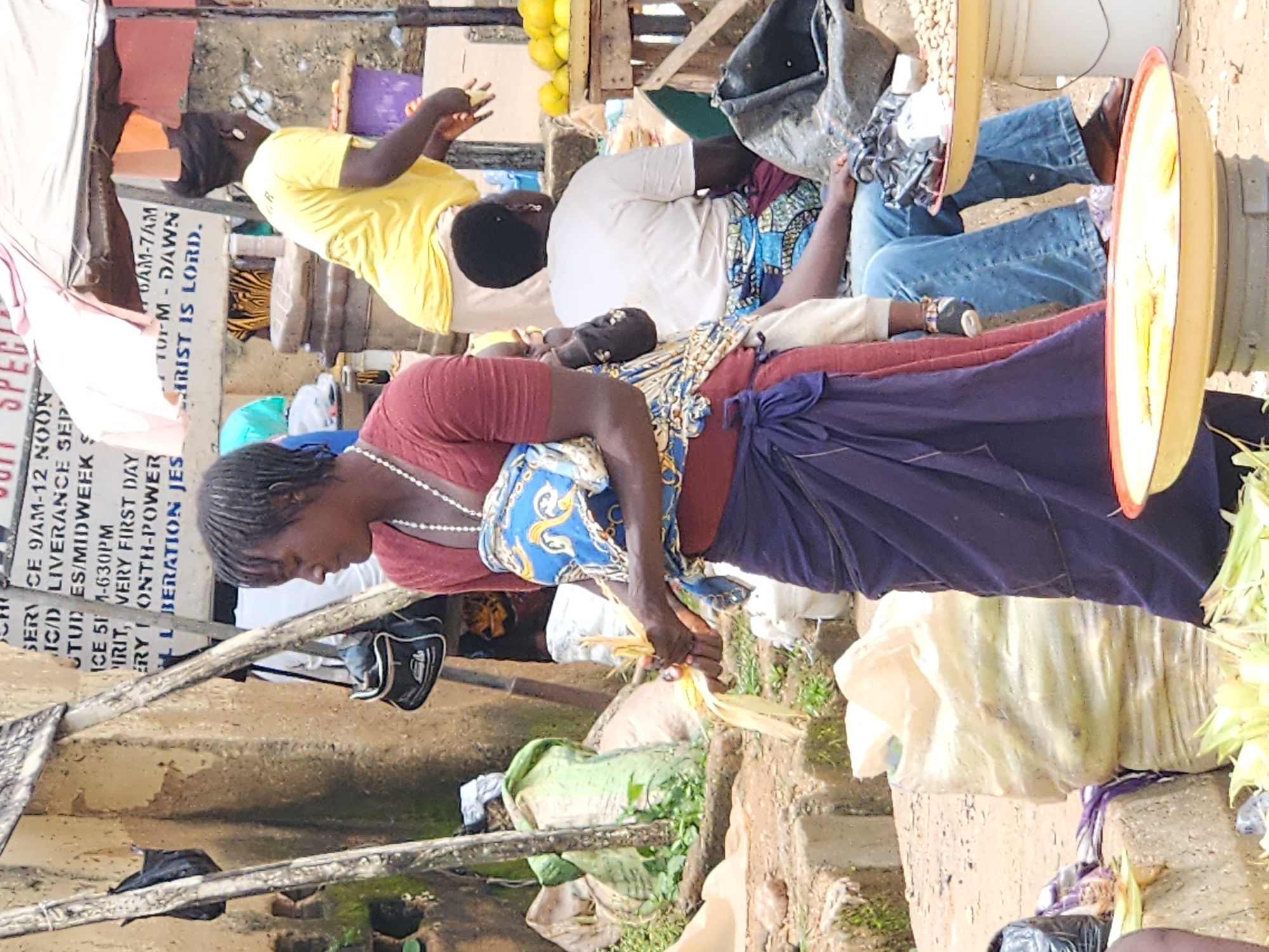 The image depicts a bustling outdoor market scene in Kabong, Nigeria. The foreground features a woman in a red shirt and a long purple skirt, carrying a baby wrapped in a blue and yellow patterned cloth. She is holding some corn husks in her hands. To her right, another woman in a yellow shirt and light-colored pants is visible, appearing to be interacting with customers or tending to her stall. In the background, a person in a white shirt sits down. The market is characterized by stalls with goods displayed on wooden tables and in bags. Items like oranges, nuts, and large sacks of produce are present. A sign with writing is visible on a wall in the background, providing context about church services. The environment is rustic, with dirt and concrete surfaces, and buildings constructed from what appears to be mud or concrete. The overall impression is one of vibrant daily life and commerce within a Nigerian urban setting.