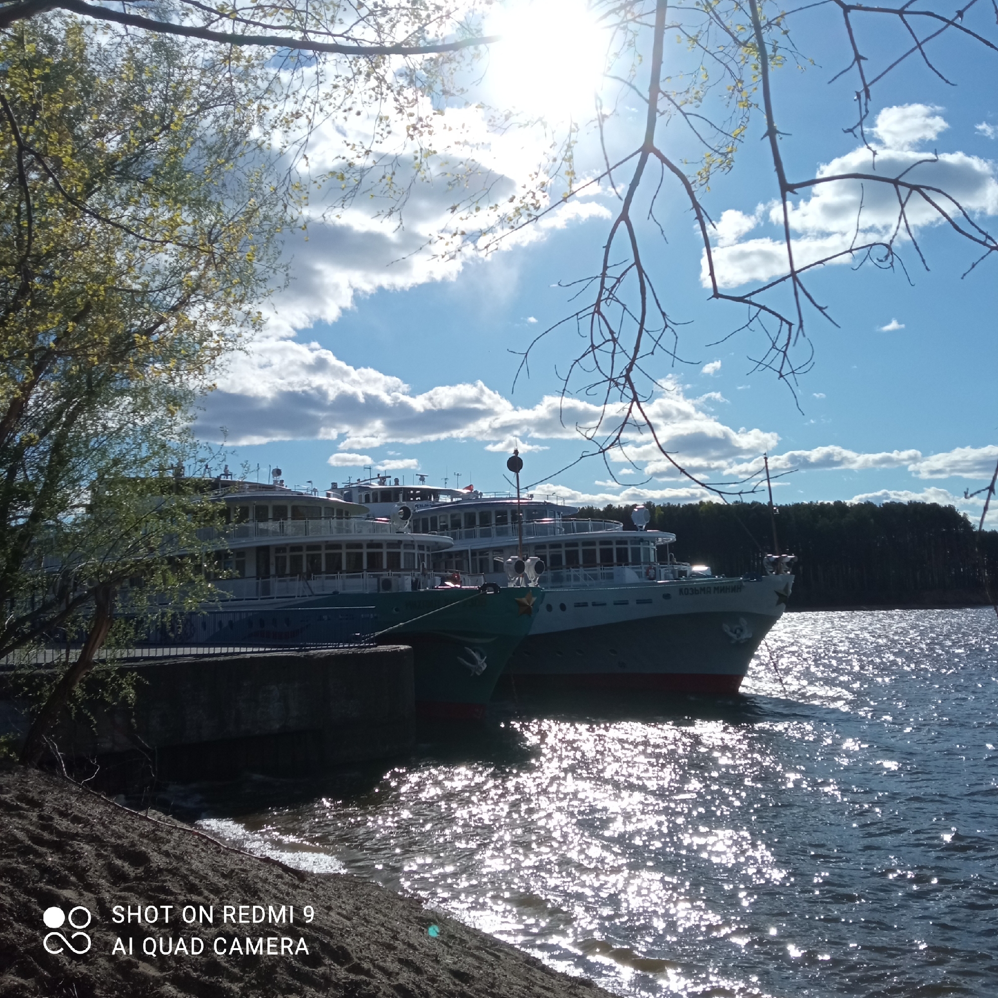 The image captures a waterfront scene in Chaikovsky, Russia. Several large passenger ships are docked along a concrete pier. The ship on the right is named "Kozma Minin." The water is reflective, indicating sunlight. The shoreline consists of an embankment of dirt or sand. There are trees in the background and along the left edge of the image, as well as bare branches in the top right. The sky is blue with scattered clouds, and the sun is visible near the top. The image was taken with a Redmi 9 AI Quad Camera, according to the watermark.