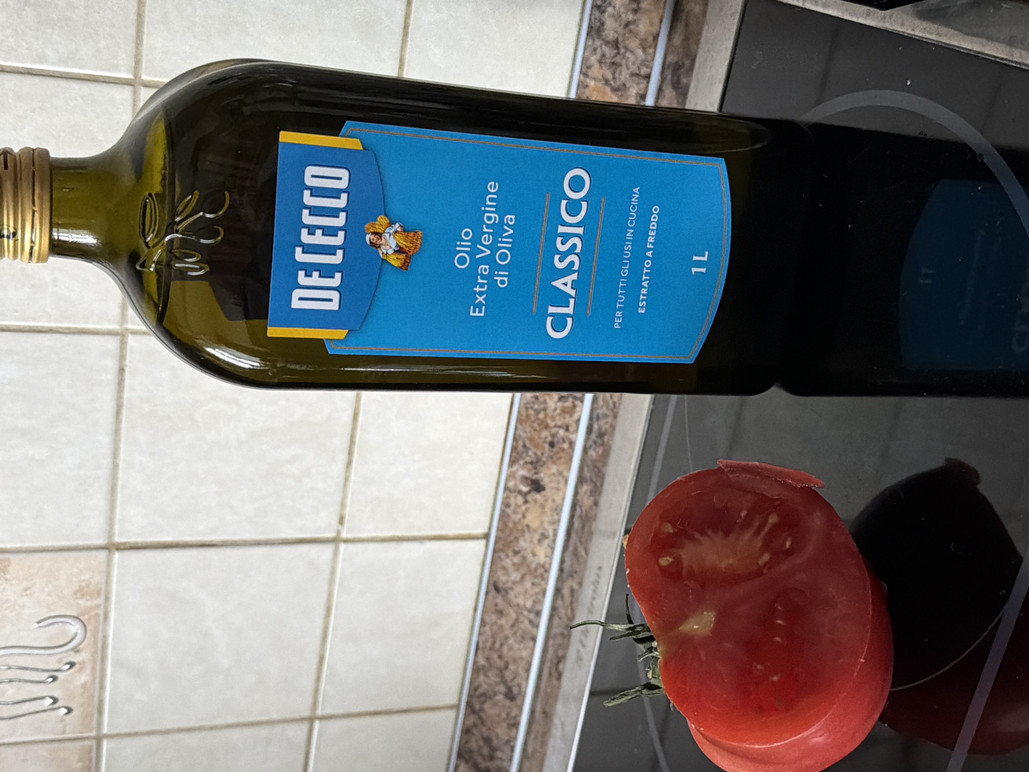 The image shows a bottle of De Cecco Classico Extra Virgin Olive Oil lying horizontally on a surface, likely a kitchen counter or stovetop, with half of a tomato next to it. The olive oil bottle is dark green glass with a gold cap. The label is blue with white lettering, indicating it's "Classico" extra virgin olive oil. The tomato appears freshly cut, with a red interior and green stem visible. The background includes light-colored tiles and a dark stovetop surface, with some kitchen utensils or accessories visible in the periphery. There's no indication of a specific location such as Khimki, Russia, within the image itself.