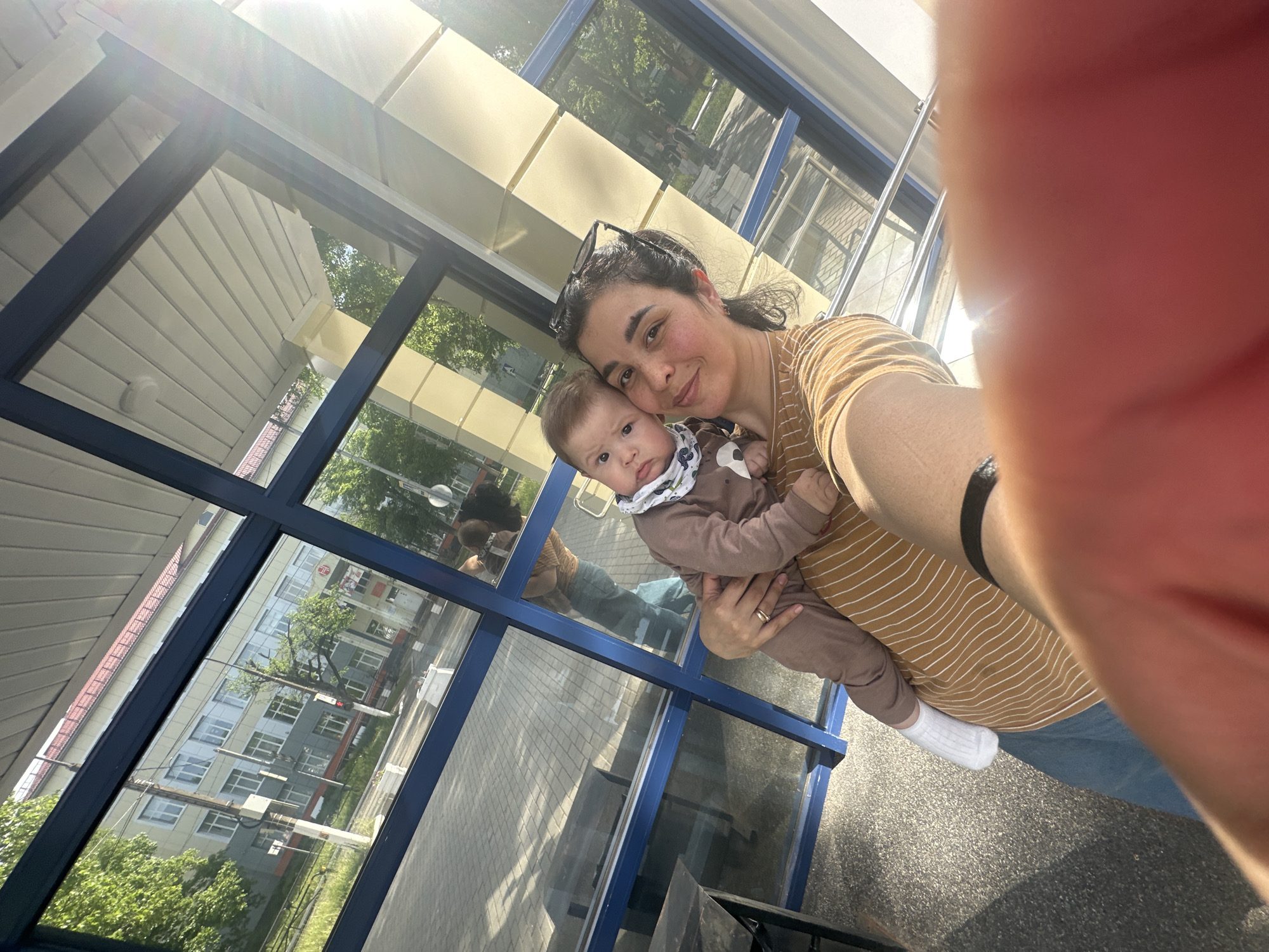 A woman is holding a baby in front of a window. The woman has dark hair pulled back, wearing a yellow striped shirt, and eyeglasses on her head. The baby is wearing a brown onesie with a white bib. They are standing next to a large window with blue frames, reflecting the buildings and trees outside. The location is unknown. The lighting suggests it is daytime. A portion of the woman's hand is visible in the foreground, likely holding the camera.