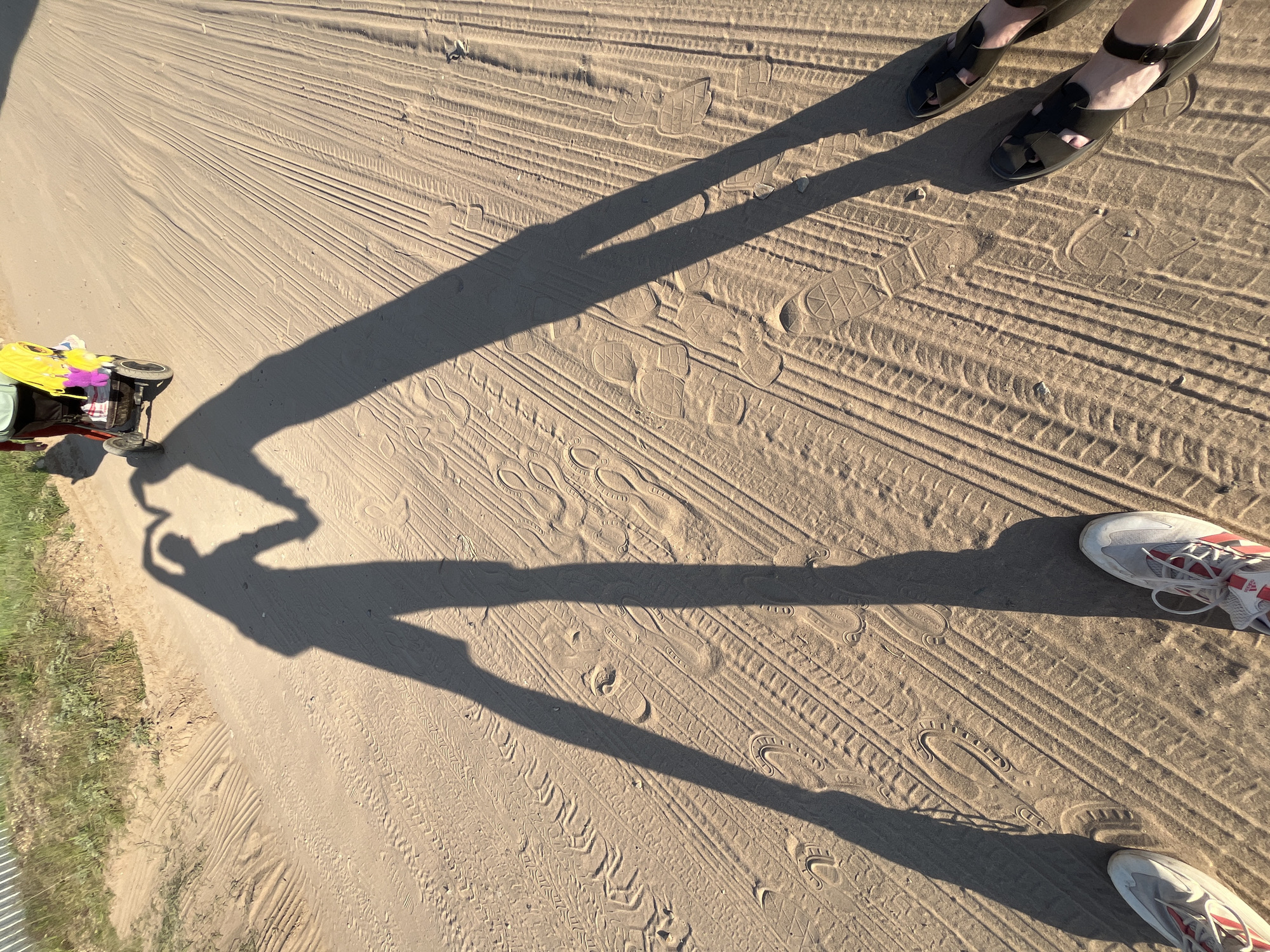The image shows a sandy area, possibly a beach or desert-like landscape. Visible are tire tracks and footprints imprinted in the sand. Shadows of two people and what appears to be a baby stroller or carriage are cast on the surface. One person is wearing sandals and the other is wearing athletic shoes. The location is believed to be in Chaikovsky, Russia based on the provided context. A small amount of grass and vegetation is seen to the left.