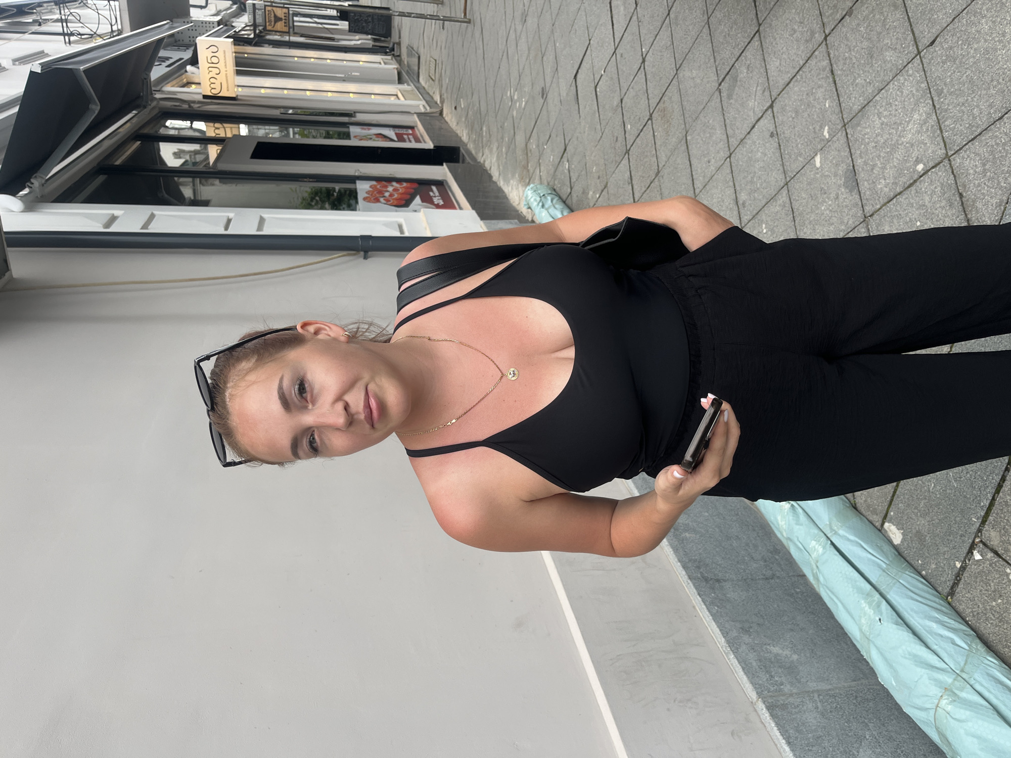 A young woman stands on a tiled sidewalk in front of a building in Batumi, Georgia, holding a smartphone in her right hand. She is wearing a black tank top, black pants, and sunglasses perched on her head. A black handbag is slung over her left shoulder. Her skin has a visible tan line on her right shoulder. The building behind her features storefronts with awnings and signage, one of which displays Georgian script. To her left, a pale blue, rolled-up object is visible on the sidewalk. The overall scene suggests an urban outdoor environment.