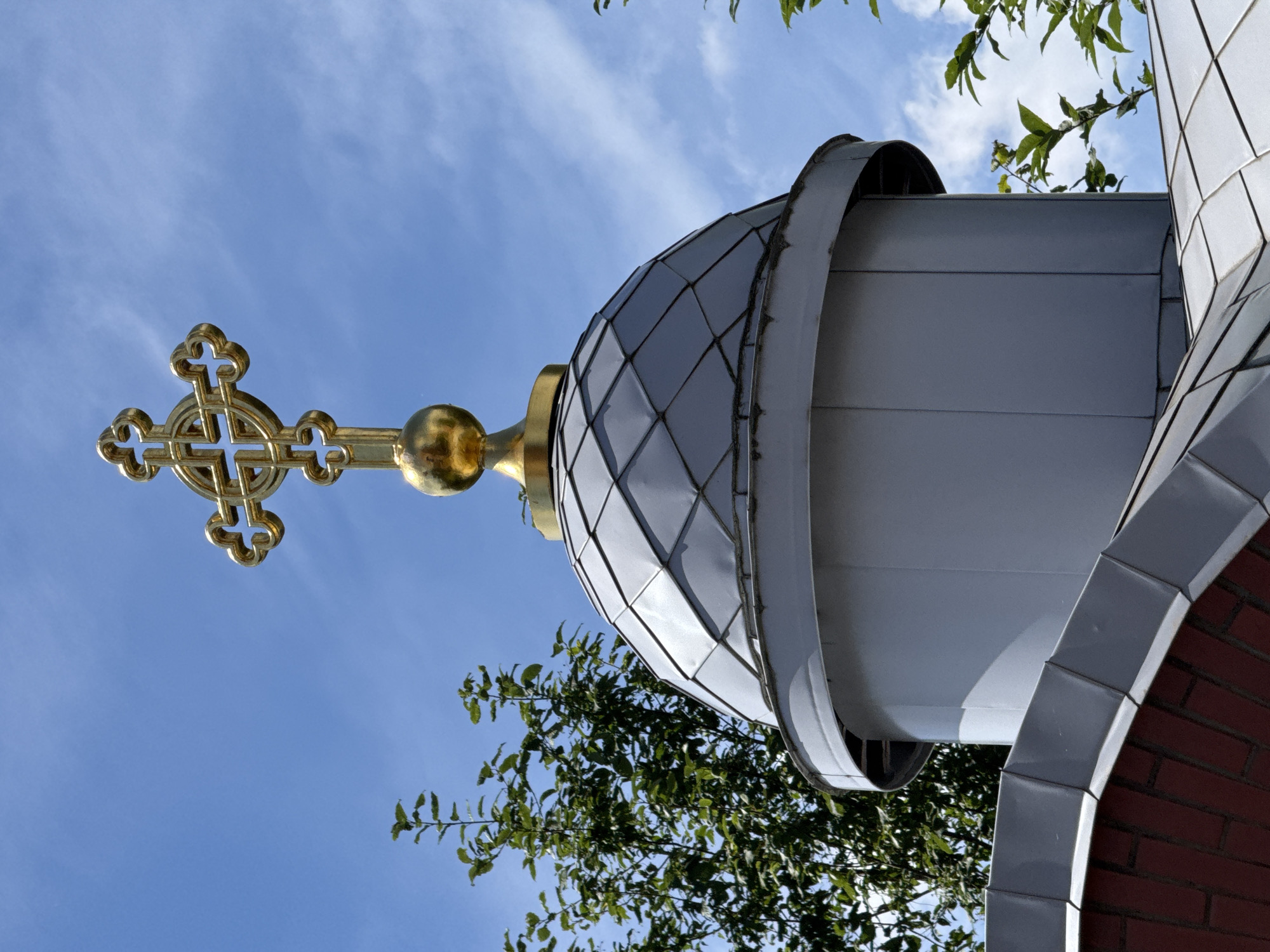 The image is a low-angle shot of the golden cross and dome of a church or cathedral, set against a bright blue sky with wispy clouds. The dome is clad in diamond-shaped metal panels, with a distinct curved edge. To the right, a section of red brick wall is visible, also adorned with metal trim. Lush green foliage from nearby trees frames the lower portion of the dome and intrudes into the frame from the top right. The golden cross, intricately designed, sits atop a golden orb, pointing upwards towards the sky. The lighting suggests it is daytime, likely sunny with fair weather. The perspective gives a sense of grandeur and architectural detail. The location context indicates this is Saint Petersburg, Russia. No people or text are visible in the image.