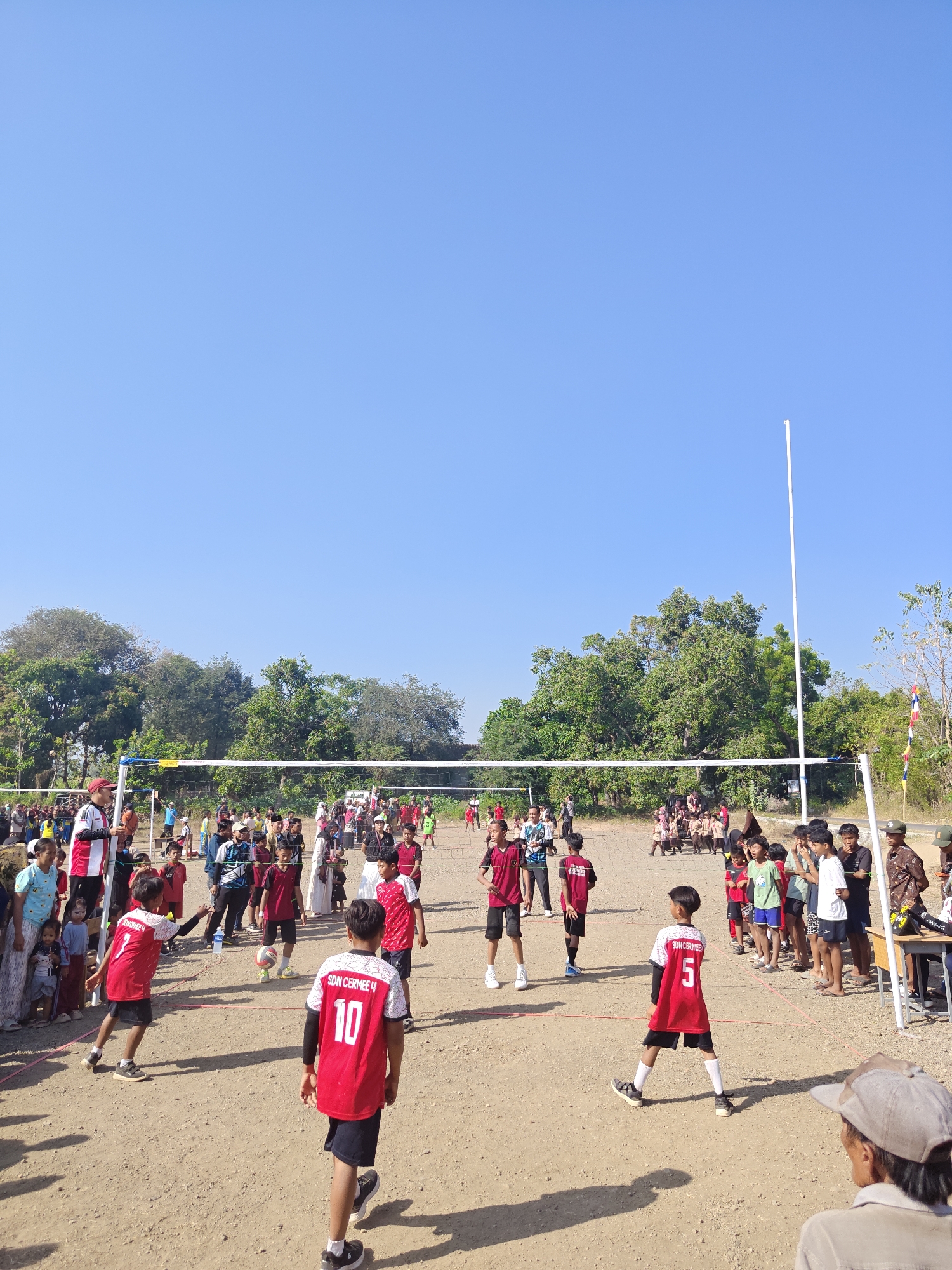 An outdoor, daytime scene depicts a volleyball game in progress, likely in the city of Cermee, Indonesia, given the visible text "SDN CERMEE 4" on several players' jerseys. The game is played on a dirt court marked with red lines. A volleyball net is stretched across the court, supported by white poles. A group of young boys, all wearing red jerseys and black shorts, are actively participating in the game. One boy in the foreground, wearing jersey number 10, faces away from the camera, while another, with jersey number 5, is in motion. A third player, with jersey number 7, is mid-action, seemingly about to hit the ball. A volleyball is visible in the air near the center of the court. Spectators, including adults and children, are gathered on both sides of the net, observing the game. Trees and foliage form a backdrop to the scene, and a clear blue sky dominates the upper portion of the image. A white flagpole stands tall on the right side of the court.