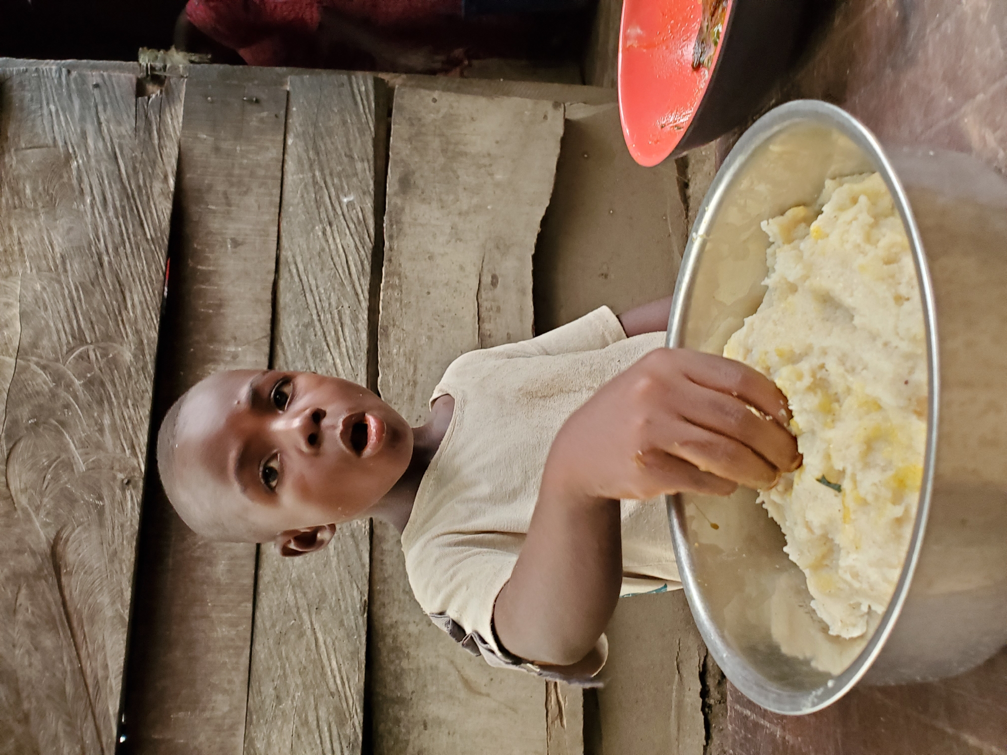 A young, dark-skinned child is reaching into a large metal bowl filled with a starchy, light-colored food, likely fufu, a staple food in Nigeria. The child is standing in a rustic setting, possibly a home, with wooden plank walls and a dirt or unfinished floor. The setting suggests the scene could be in a less affluent area of Mararaba, Nigeria. There is another bowl, red in color, partially visible in the upper right corner, suggesting that the child may be preparing to eat or is in the process of eating. The child’s open mouth and expression indicate surprise or a call for attention.