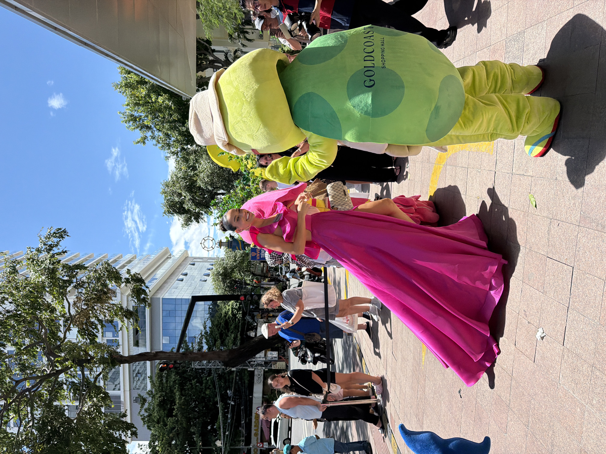 In Nha Trang, Vietnam, on a sunny day, a woman in a vibrant hot pink gown stands on a pedestrian walkway. She is smiling and looking to her left. To her right, a person in a large green turtle mascot costume, with a hat and "Goldcoast Shopping Mall" written on its back, is facing away from the camera, with its arms extended. Several other people are visible in the background, including onlookers, a person holding a camera, and others standing in line. The environment includes a street with parked motorcycles, trees, and a modern building with glass windows. A blue, fin-like object is on the ground to the left of the frame.