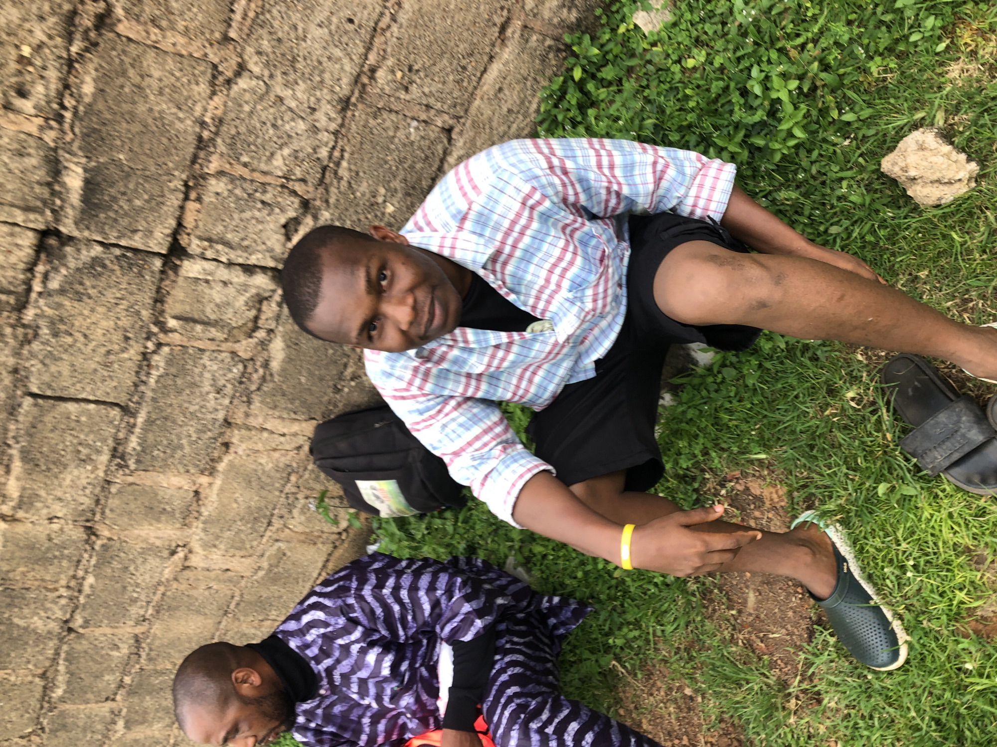 Two adult males are seated on the ground in a grassy area next to a rough, weathered brick wall. The male on the left wears a dark top with a black turtleneck and a purple and black patterned robe-like garment. He is partially obscured and is facing left. The male on the right is closer to the camera, facing forward and looking at the camera. He is wearing a plaid shirt with red and blue stripes over a black t-shirt, black shorts, and dark clogs. A yellow wristband is visible on his left wrist. A black backpack is positioned behind the man on the left, leaning against the brick wall. The ground is covered in grass with some sparse patches of dirt and small green plants. A piece of light brown, textured material, possibly dried mud or a small rock, is on the ground near the legs of the man on the right. The lighting suggests an outdoor daytime scene. The environment is consistent with an outdoor setting in Jos, Nigeria.