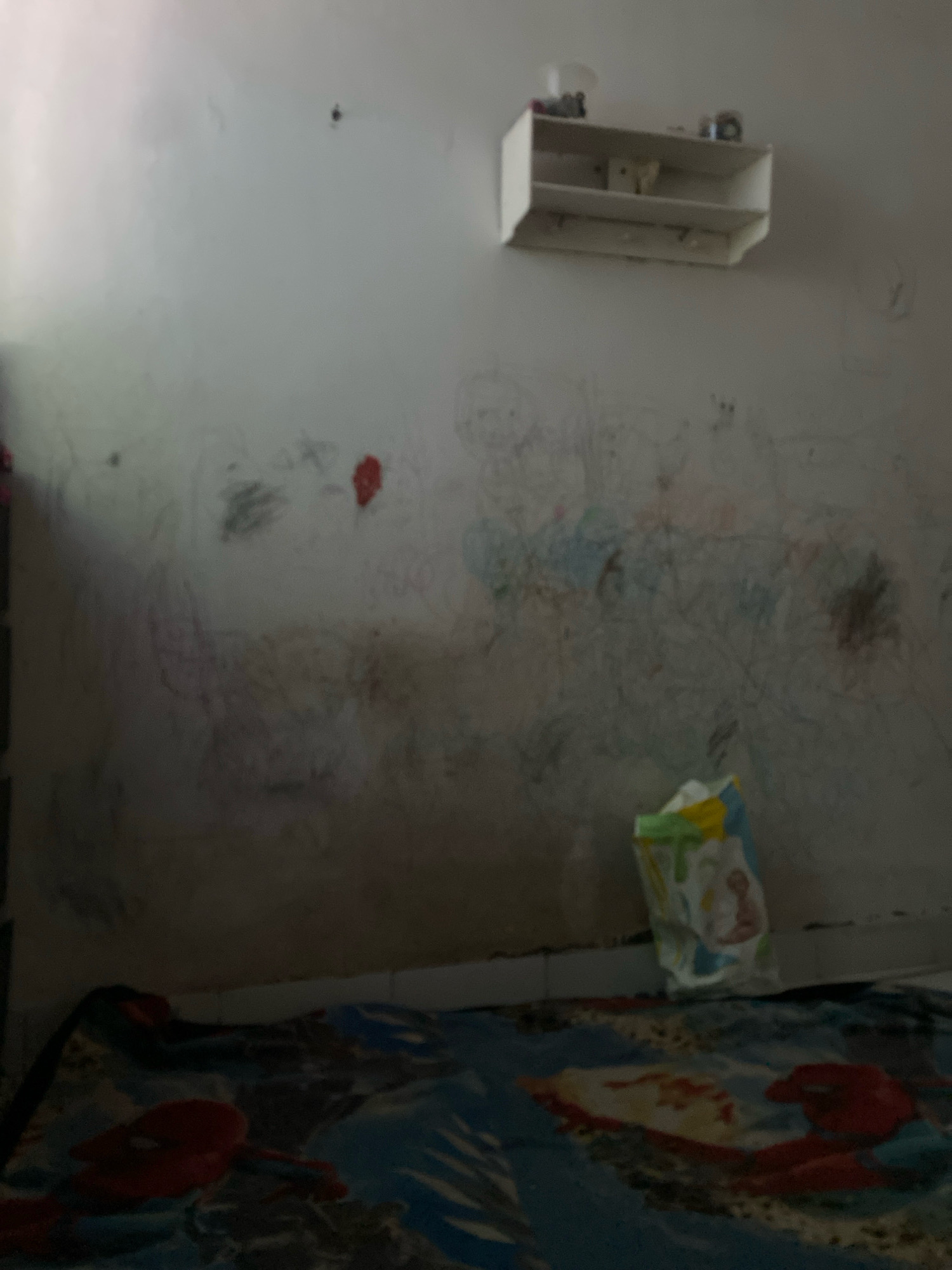 The image depicts a dimly lit room, possibly a child's bedroom, with a wall covered in various colorful scribbles and drawings, suggesting it is a place where a child has been active. A white shelf is mounted on the upper part of the wall, holding a few small, indistinct objects. Below the shelf, a crumpled bag with colorful patterns, resembling a shopping bag or a toy packaging, stands upright on what appears to be a patterned blanket or bedspread. The blanket has a prominent red and blue design, possibly featuring cartoon characters. The overall impression is one of a lived-in, somewhat messy space. The provided metadata does not contain information about the location being Bandar Lampung, Indonesia.