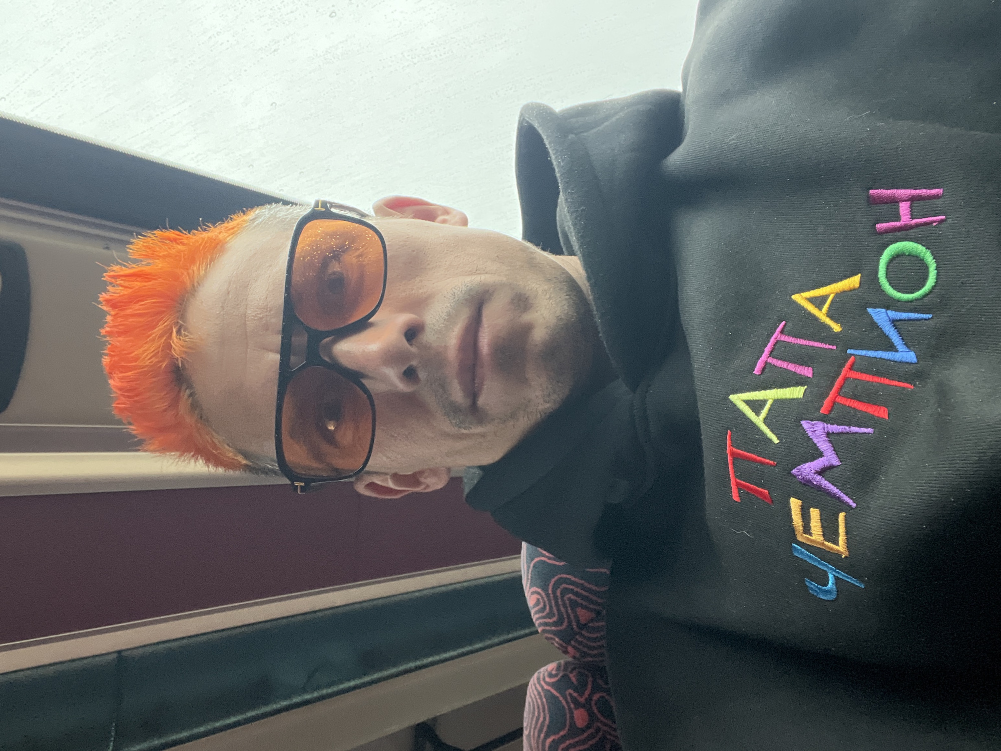 A man with bright orange hair and amber-tinted glasses is inside what appears to be a train car, likely on a rainy day based on the water droplets on the glasses and the window above him. He is wearing a black hooded sweatshirt with colorful embroidery in Cyrillic script that translates to "Papa Champion." The interior of the train car shows a maroon panel, a metallic or light-colored rail, and a darker panel below. The man is looking directly at the camera. The train car suggests the location could be in Chaikovsky, Russia.