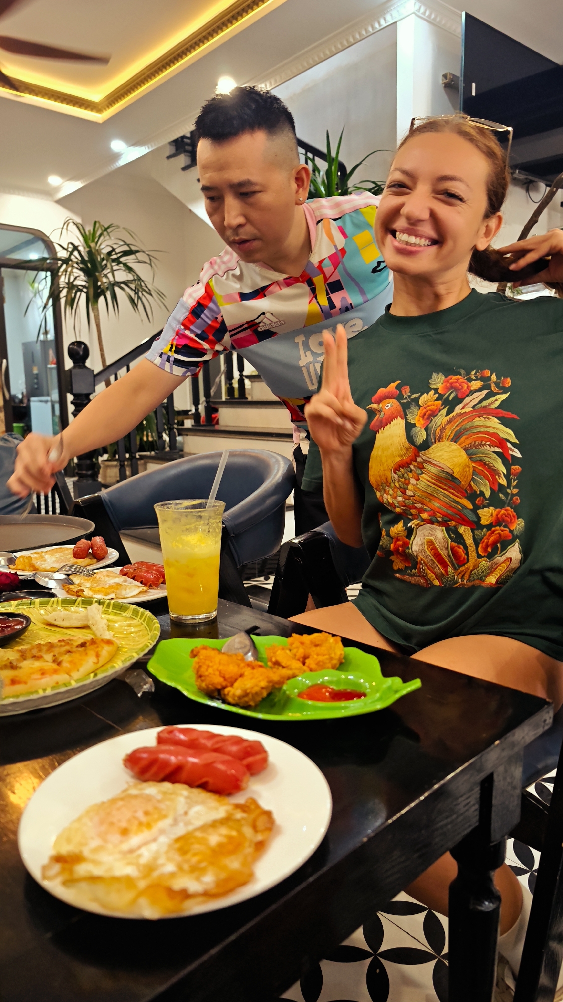 The image captures two people, a man and a woman, enjoying a meal at an indoor restaurant or cafe, likely in Phố Ràng, Vietnam. The woman, positioned in the foreground and to the right, is smiling brightly at the camera, holding up two fingers in a "V" sign with her right hand. She wears a dark green t-shirt adorned with a large, colorful rooster and floral design. Her left hand is near her light brown hair, and she has glasses resting on top of her head, expressing a cheerful and happy emotion.
To the left and slightly behind her is a man with short, dark hair, wearing a vibrant, multi-colored short-sleeved shirt with an Adidas logo on the sleeve and "LOVE U" text partially visible on the front. He appears focused on the food, leaning over the table with a fork in his hand, as if serving or about to eat.
The black wooden table in front of them is laden with various dishes. On a white plate are two fried eggs and two bright red sausages. A bright green, leaf-shaped plate holds several pieces of fried chicken next to a small dish of red sauce. Another yellow, patterned plate contains a pizza-like dish with some slices already eaten. A tall glass of yellow juice with a straw is also on the table.
The background reveals a well-lit interior with a grand staircase featuring a dark banister. Green potted plants are visible near the stairs. The ceiling has decorative gold-colored molding and recessed lighting, and a ceiling fan is partially visible. The floor is covered with a distinctive black and white geometric tiled pattern, suggesting a stylish setting. The scene depicts a lively and casual dining experience.