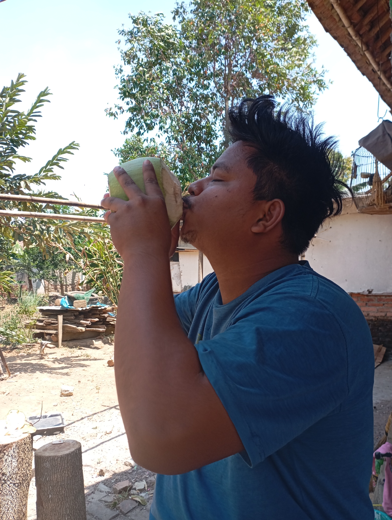 An Indonesian male with dark, spiky hair is shown from the chest up, drinking from a light green gourd. He is wearing a blue t-shirt. The background appears to be an outdoor, rural setting with trees, a wooden structure with a thatched roof, and a birdcage. The ground is dry and dusty, with scattered rocks and some vegetation. The sky is bright blue and clear. The image appears to be taken in Kecamatan Haurgeulis, Indonesia, based on the context provided.