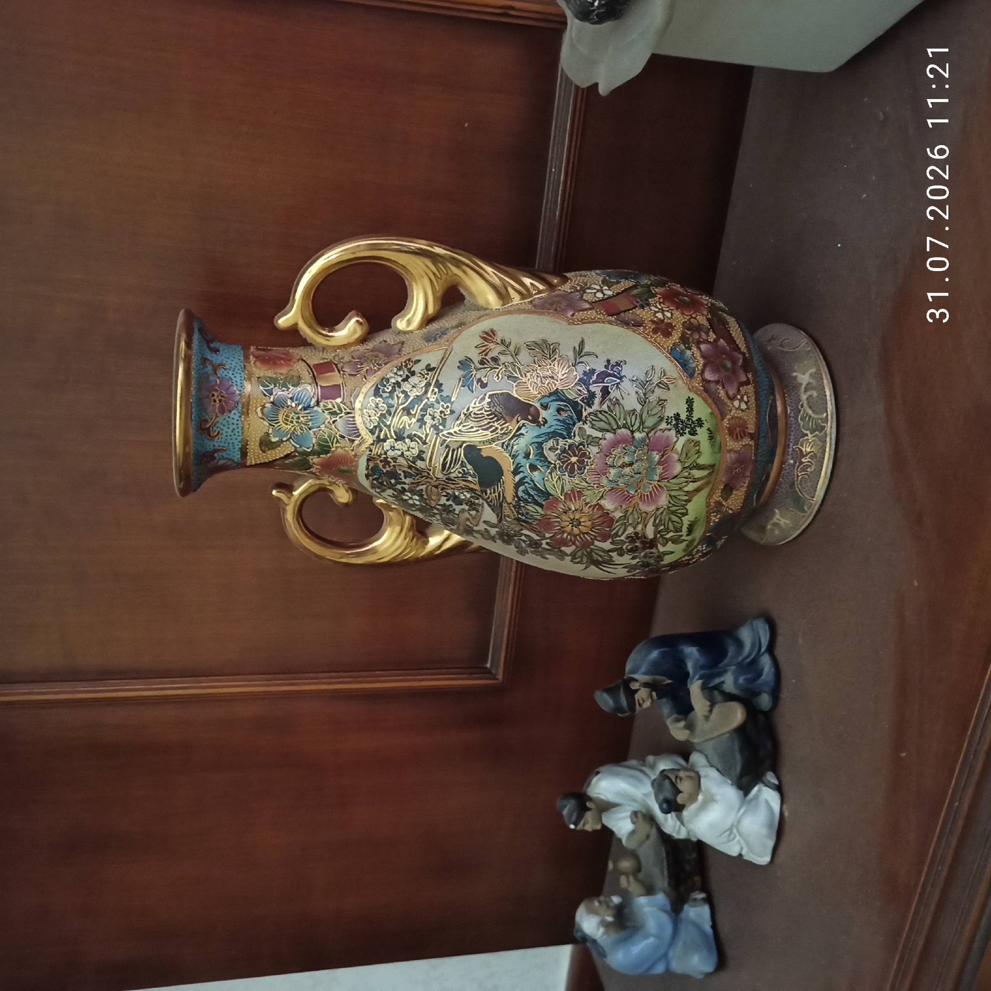 In the city of Modugno, Italy, on a wooden shelf, rests an ornate vase adorned with intricate floral and avian designs in vibrant colors. Two golden handles curve from its sides. To the left of the vase, a group of four small figurines depict men in traditional attire. One elderly figure with a white beard sits facing another man, both engaged with a small table. A third figure in white kneels beside a rock, while a fourth figure in blue is crouched, appearing to work on something. The background is a dark wooden surface, likely a cabinet or wall panel. A white object, possibly a bust or lamp base, is partially visible to the right of the vase. The timestamp "31.07.2026 11:21" is visible in the lower right corner of the image.