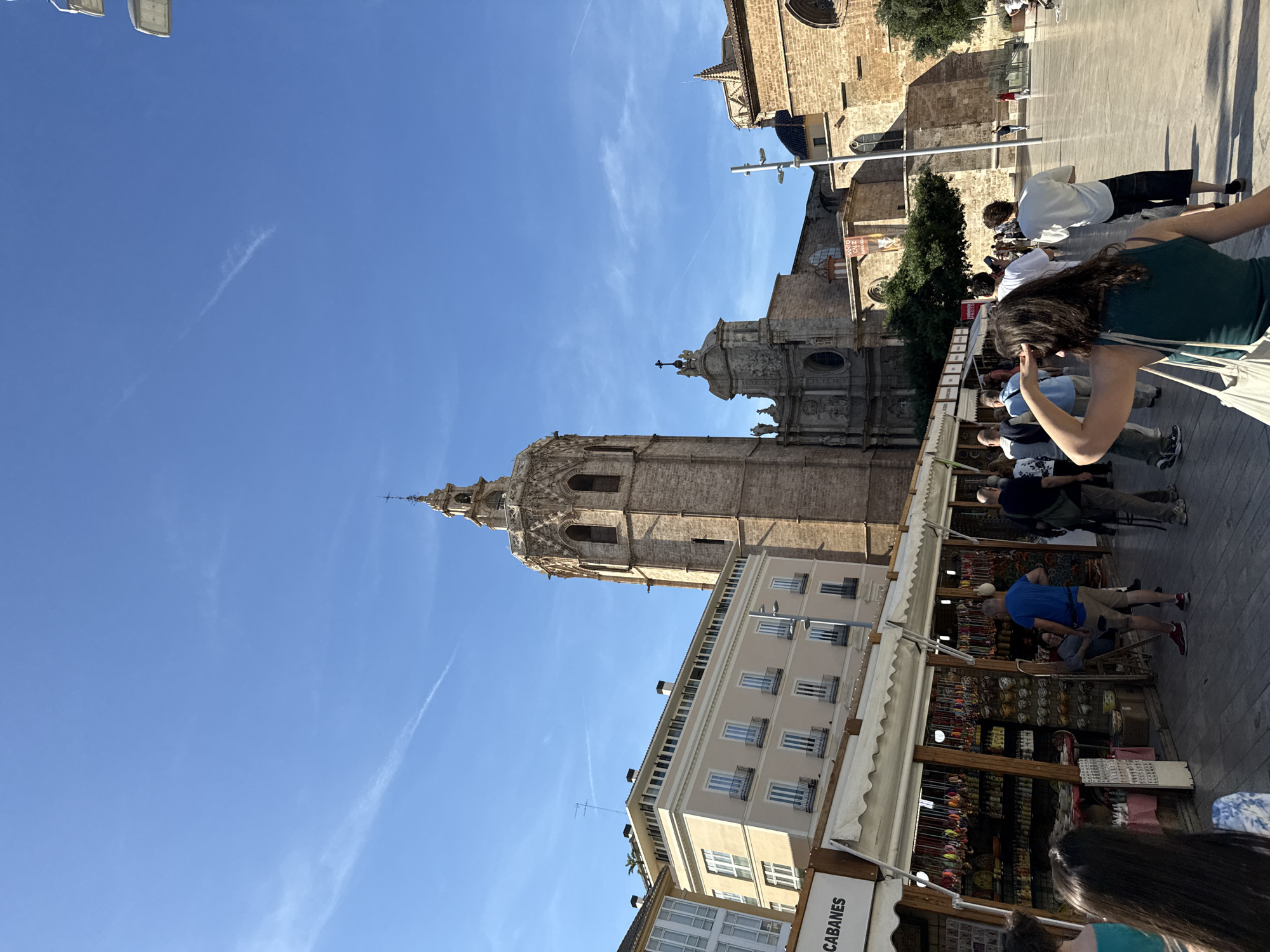 The scene depicts a bustling square in Valencia, Spain, likely near the Valencia Cathedral based on the prominent, ornate bell tower in the frame. The tower is made of light-colored stone and stands tall, piercing the clear blue sky. A row of market stalls is visible, stocked with various items and shoppers browsing. People are walking around, some appearing to be tourists taking photos. The buildings surrounding the square have architectural details, including balconies and decorative elements. The light suggests it is daytime.