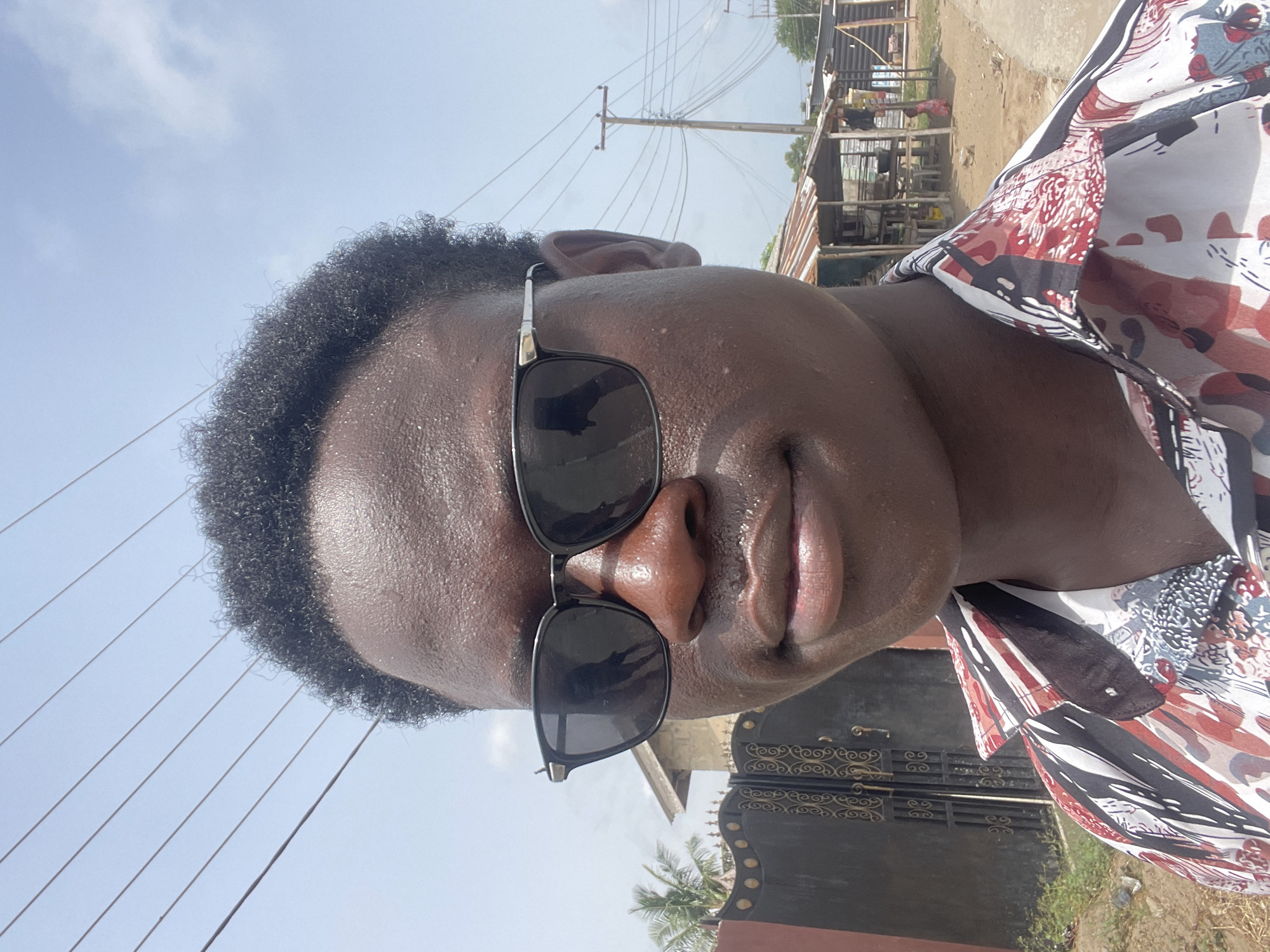 A close-up, rotated portrait shows a dark-skinned individual wearing sunglasses and a patterned shirt. In the background, utility poles with wires are visible against a light blue sky. Also present is a building with a corrugated metal roof and an ornate, wrought-iron gate. Based on the style of architecture and the overall environment, the location is consistent with a typical street scene in Badagry, Nigeria.