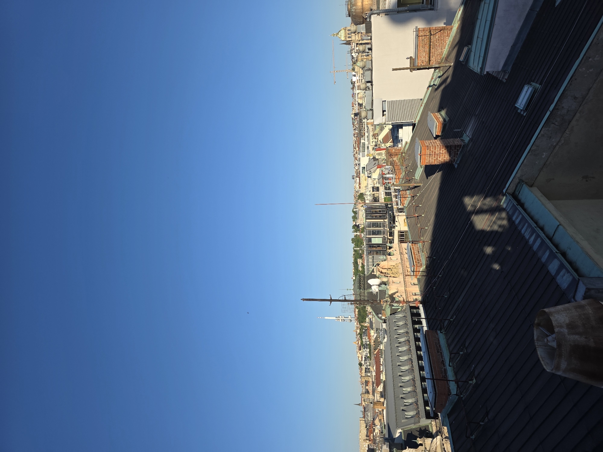 A high-angle, rotated view shows a city skyline under a clear blue sky. Dominating the foreground is a dark, sloped roof with architectural details. Beyond the roof, a dense cityscape unfolds with buildings of varying heights and styles. Towers, domes, and cranes punctuate the skyline. The city is likely located in an Unknown location. The sky is vast and cloudless. The image suggests a rooftop perspective overlooking the city.