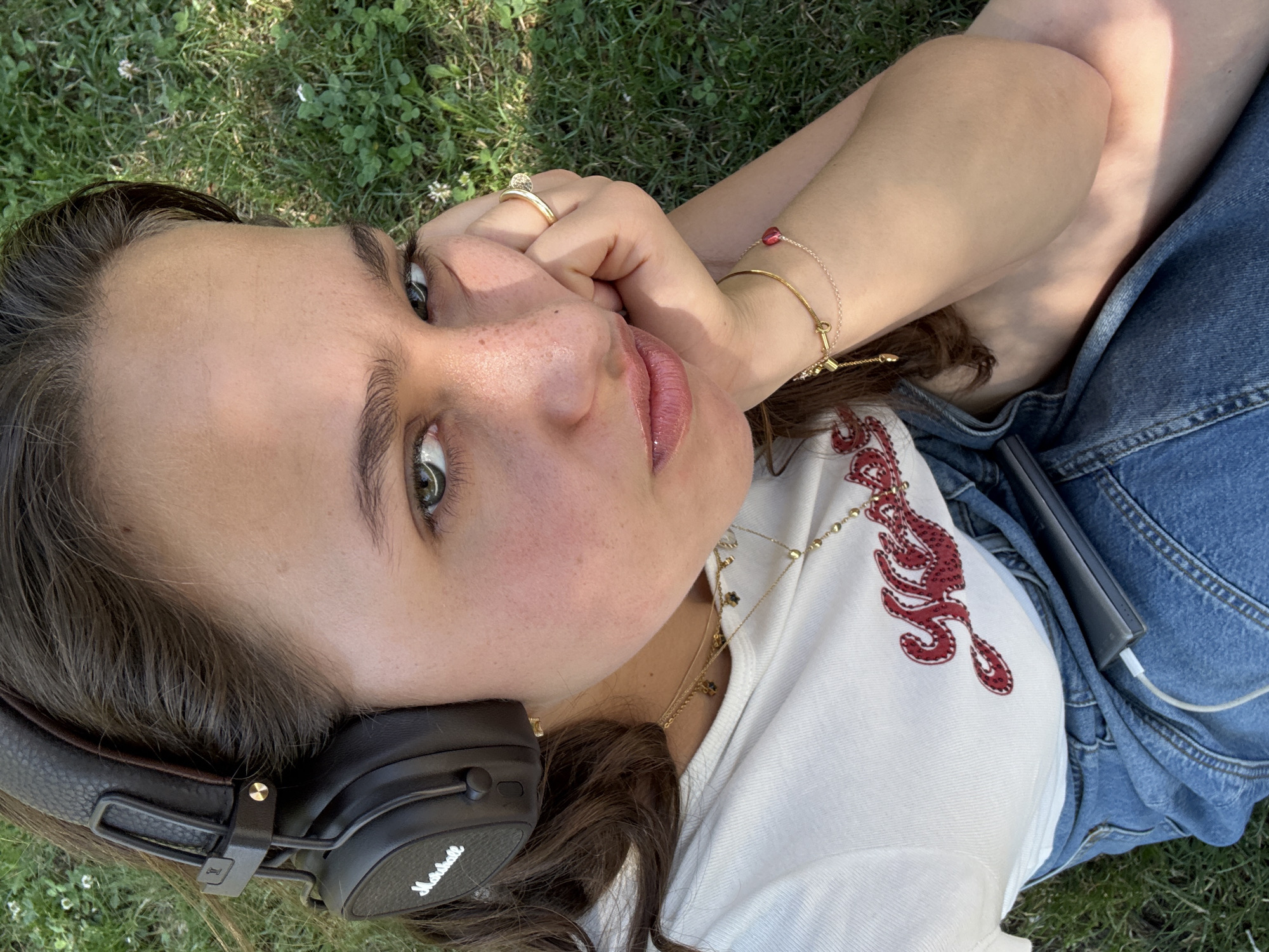 A young woman is lying on grass, wearing black Marshall headphones, a white t-shirt with a red design, and denim overalls. She is resting her head on her hand, and her expression appears thoughtful or melancholic. A power bank is connected to a white cable in her lap. The scene suggests a casual outdoor setting, possibly a park. No specific Milanese landmarks are visible.