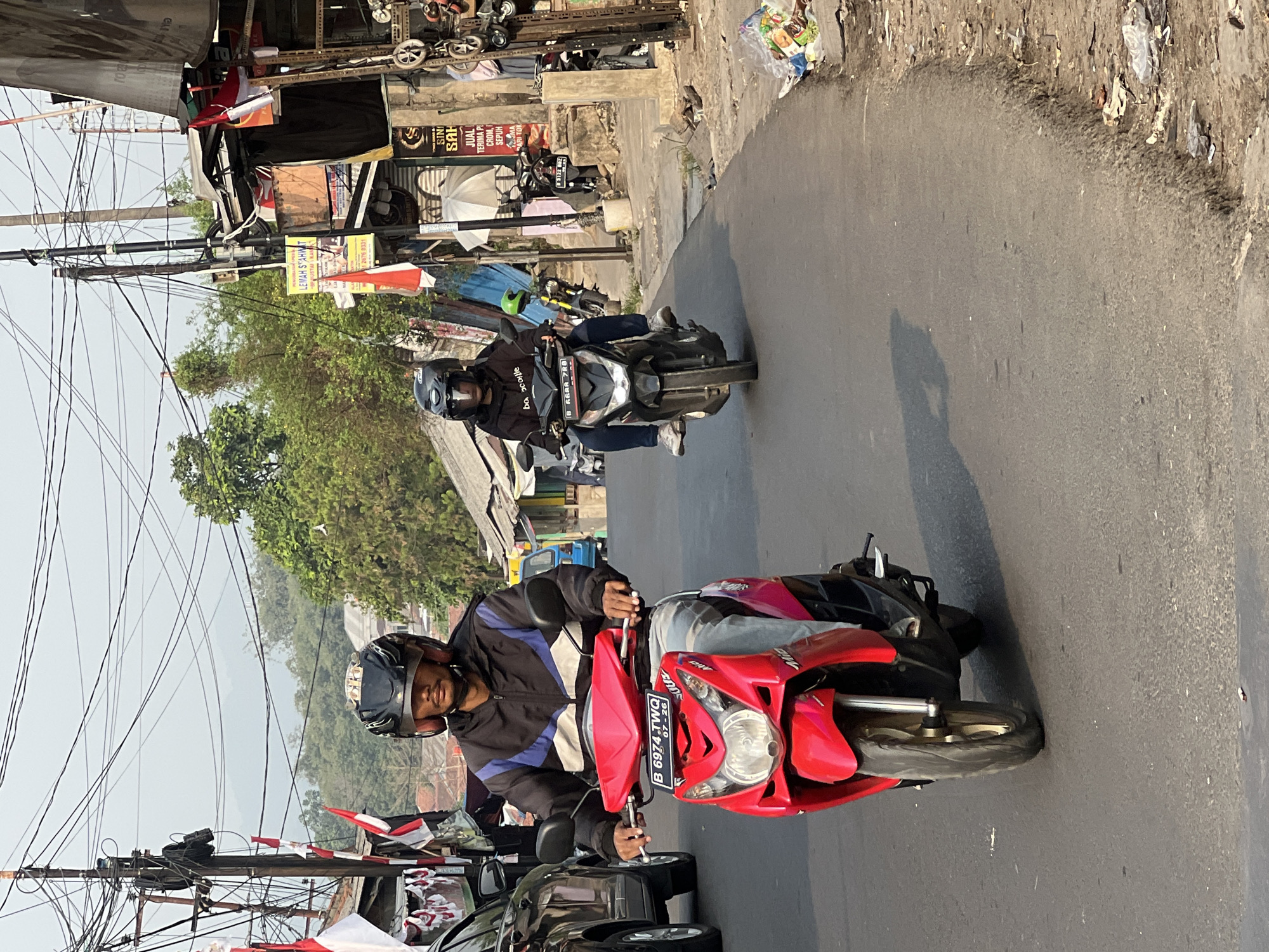 The image shows a street scene in Citeureup, Indonesia. Two individuals are riding motorcycles on a paved road. The motorcycle in the foreground is red, and the rider is wearing a black and blue jacket and a black helmet. The motorcycle in the background is black, and the rider is wearing a black hoodie and a black helmet. The road is lined with buildings and vegetation. There are utility poles with numerous wires visible. The sky is overcast. The scene suggests a typical day in an urban or semi-urban area.