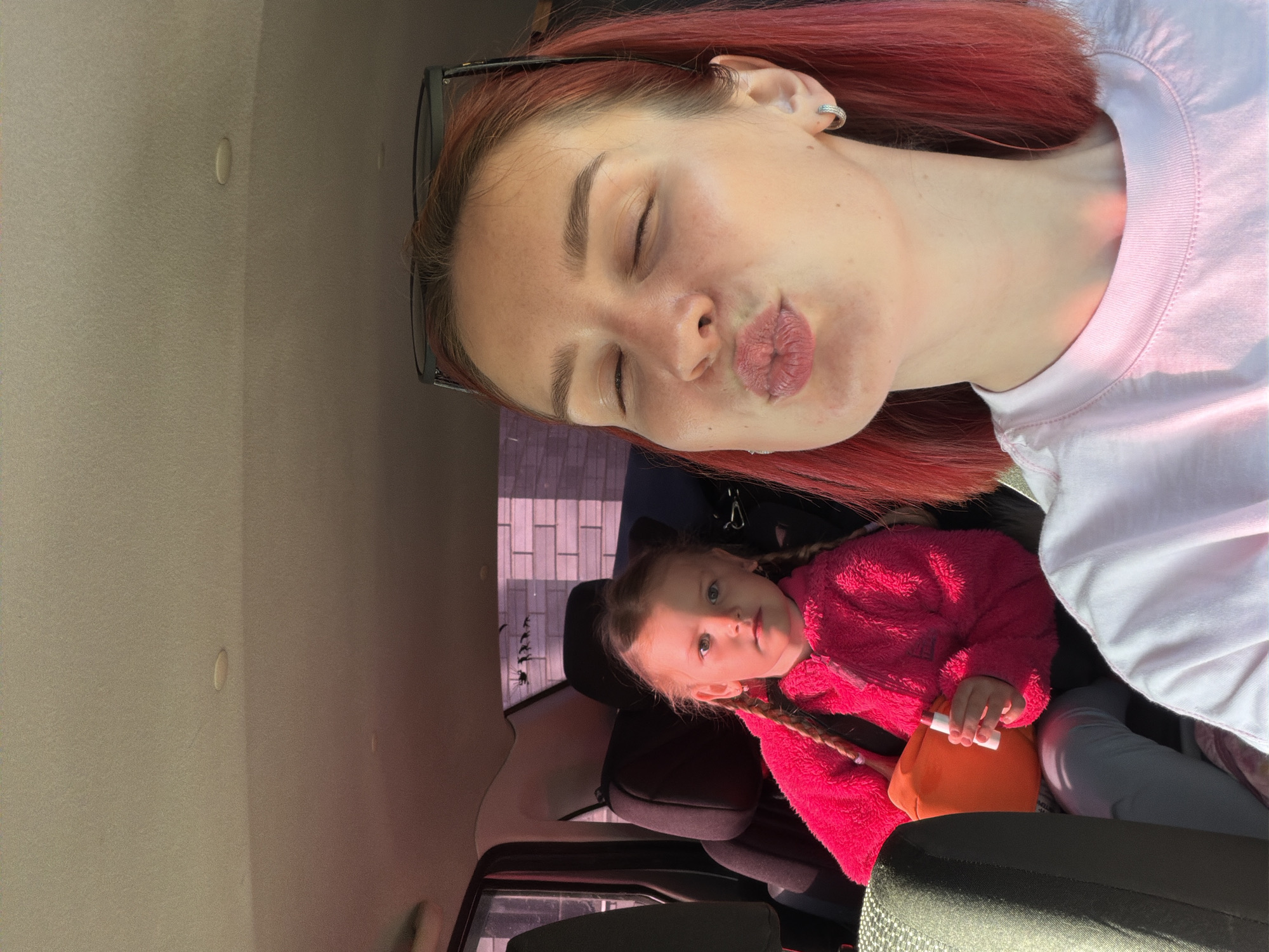 A selfie taken inside a car shows a woman with red hair and a white t-shirt puckering her lips. Behind her, a young girl with blonde hair in braids is sitting in a car seat, wearing a pink sweater and holding a white and orange object. The interior of the car is visible, with the ceiling and seats in neutral tones. A glimpse of an exterior brick wall is visible through a window in the background, implying an urban or suburban setting. The image was taken in Chaikovsky, Russia.