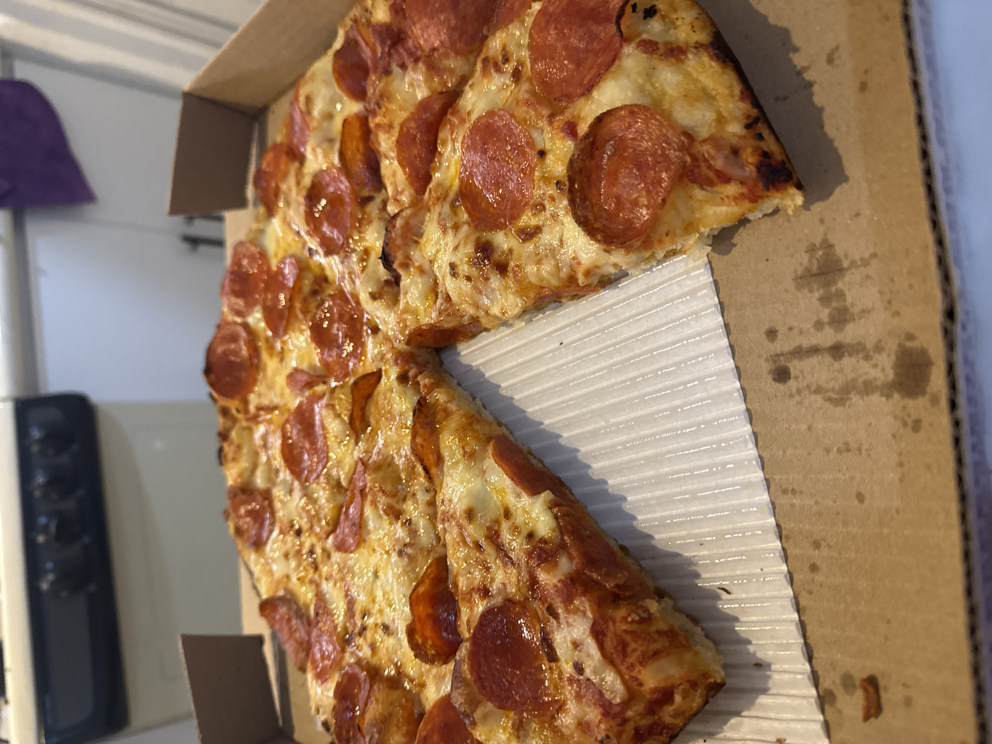 The image shows a partially eaten pepperoni pizza inside a cardboard pizza box. The pizza appears to have a thin crust and is topped with melted cheese and pepperoni slices. The box is stained with grease. The location is not discernible from the image.