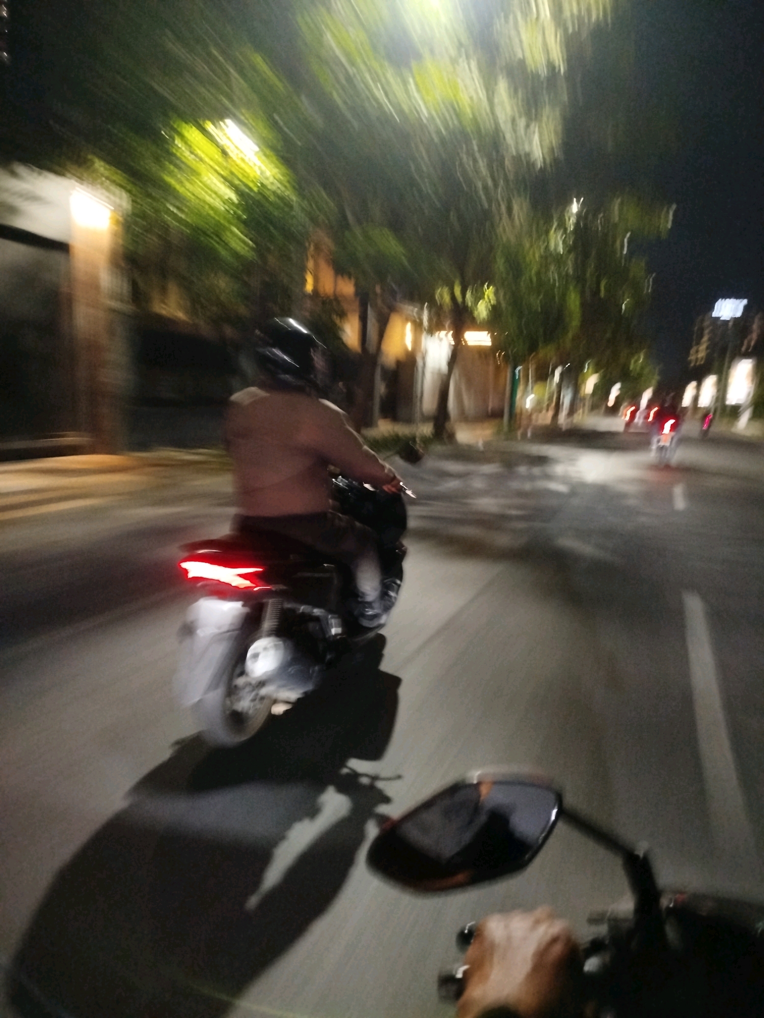 The image captures a nighttime scene on a street in Surabaya, Indonesia. The foreground shows a motorcycle's side mirror, a hand gripping the handlebar, and a portion of the motorcycle's body with a prominent shadow cast on the road. The main subject is a person riding a motorcycle ahead, illuminated by streetlights and the motorcycle's tail light. This rider is wearing a helmet and is shirtless, with their back to the camera. The background reveals trees with illuminated foliage, buildings lining the street, and other motorcycles in the distance, also with their tail lights visible. The motion blur suggests the image was taken from a moving vehicle, likely another motorcycle, on a street with moderate urban activity at night.