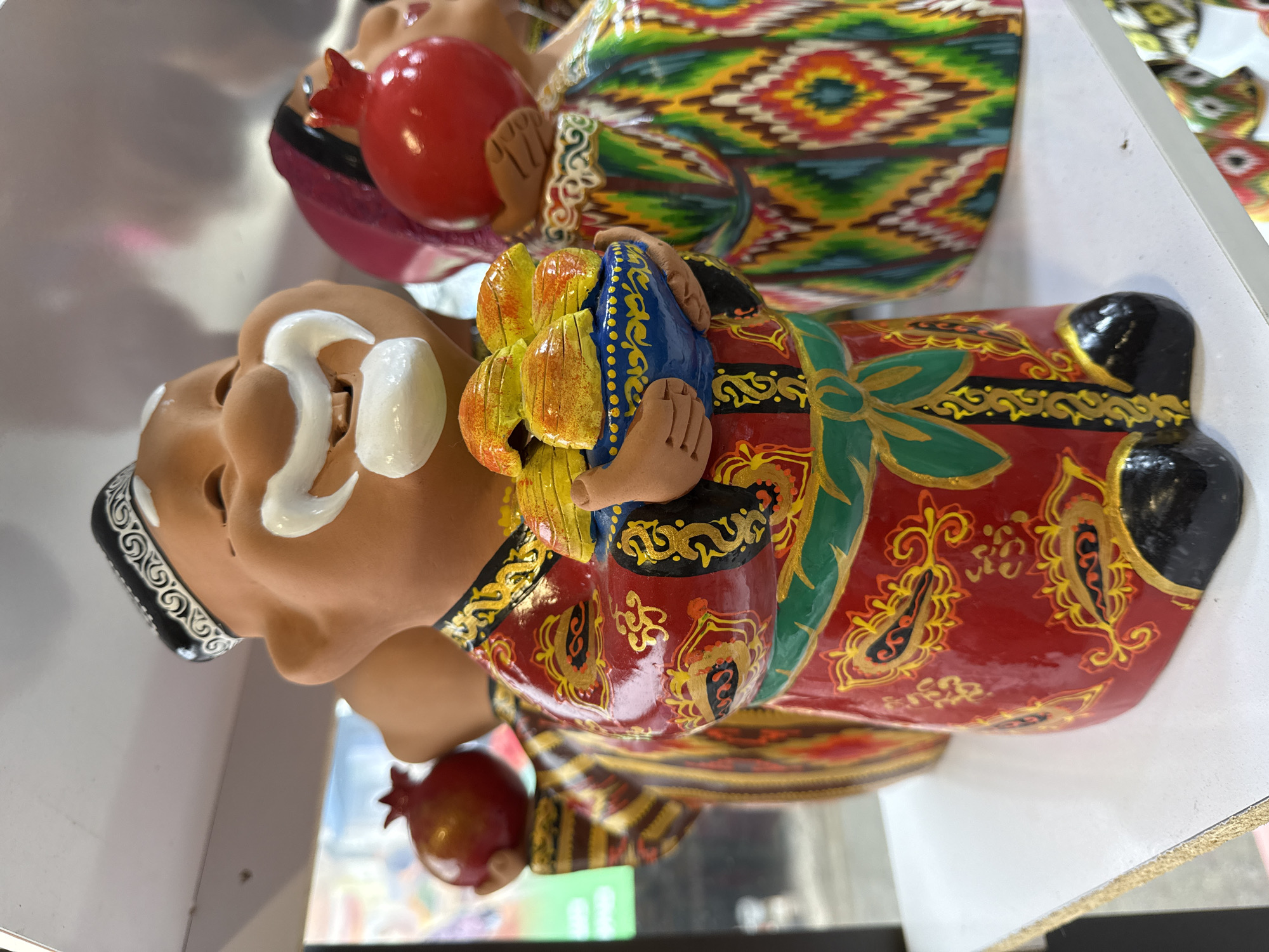 The image shows two souvenir figurines of elderly men in traditional Uzbek clothing, likely photographed in Tashkent, Uzbekistan. The figurines are brightly colored and depict men with elaborate white mustaches and beards. One figure wears a red and gold patterned robe, a green sash, and a black skullcap with white embroidery. He holds a yellow and blue object resembling a stylized flower or fruit. The other figurine wears a multi-colored patterned robe and holds a red pomegranate. Both figurines are positioned on a white surface, possibly a shelf.