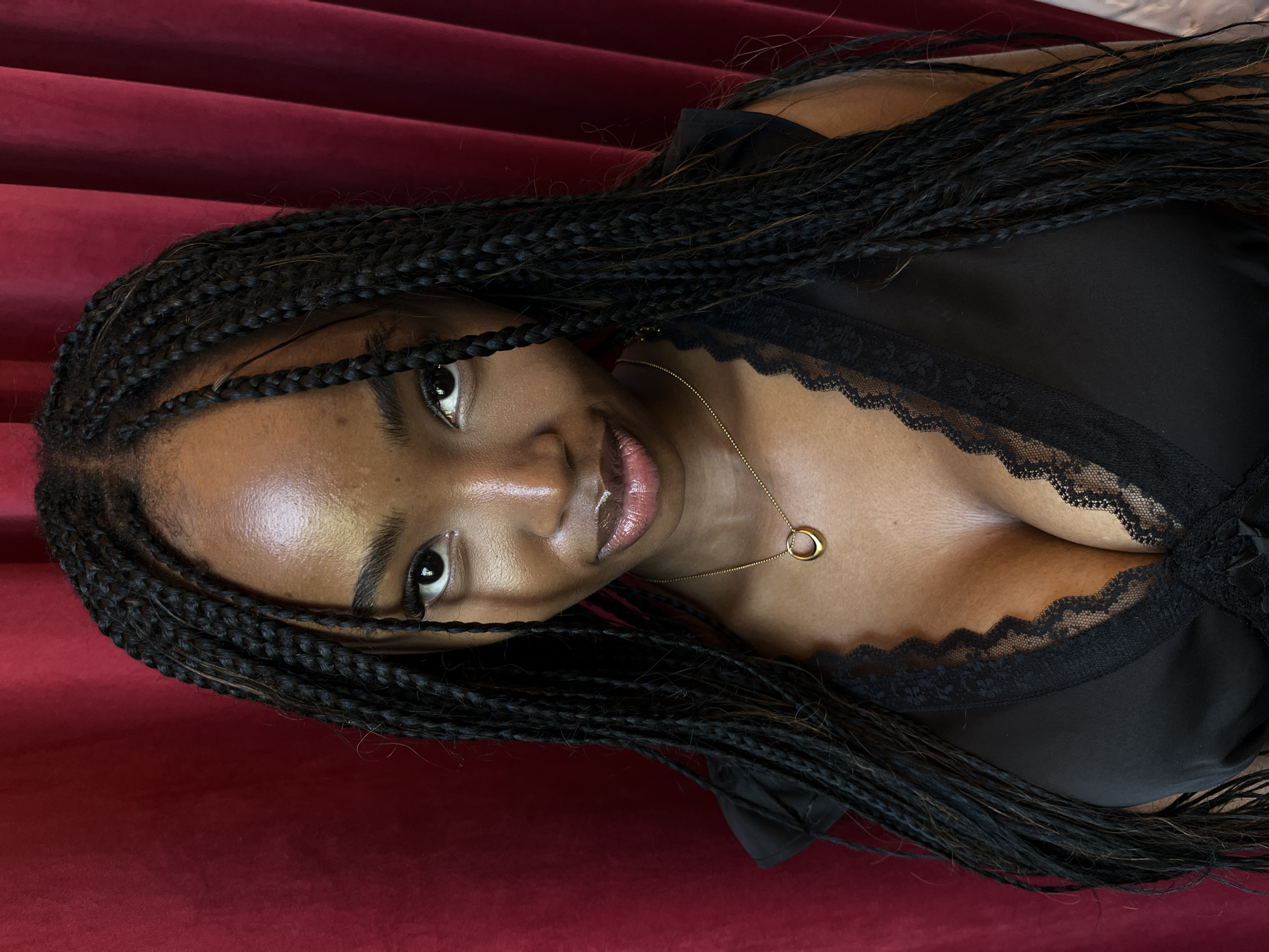 A medium shot of a young Black woman with braided hair, wearing a black low-cut top and a gold necklace, is featured against a red velvet curtain background. The woman's head is slightly tilted, and she is looking directly at the camera with a subtle smile. The lighting highlights her features, particularly her eyes and lips, and gives her skin a luminous glow. The red curtain creates a dramatic backdrop, with vertical folds adding texture and depth to the scene.