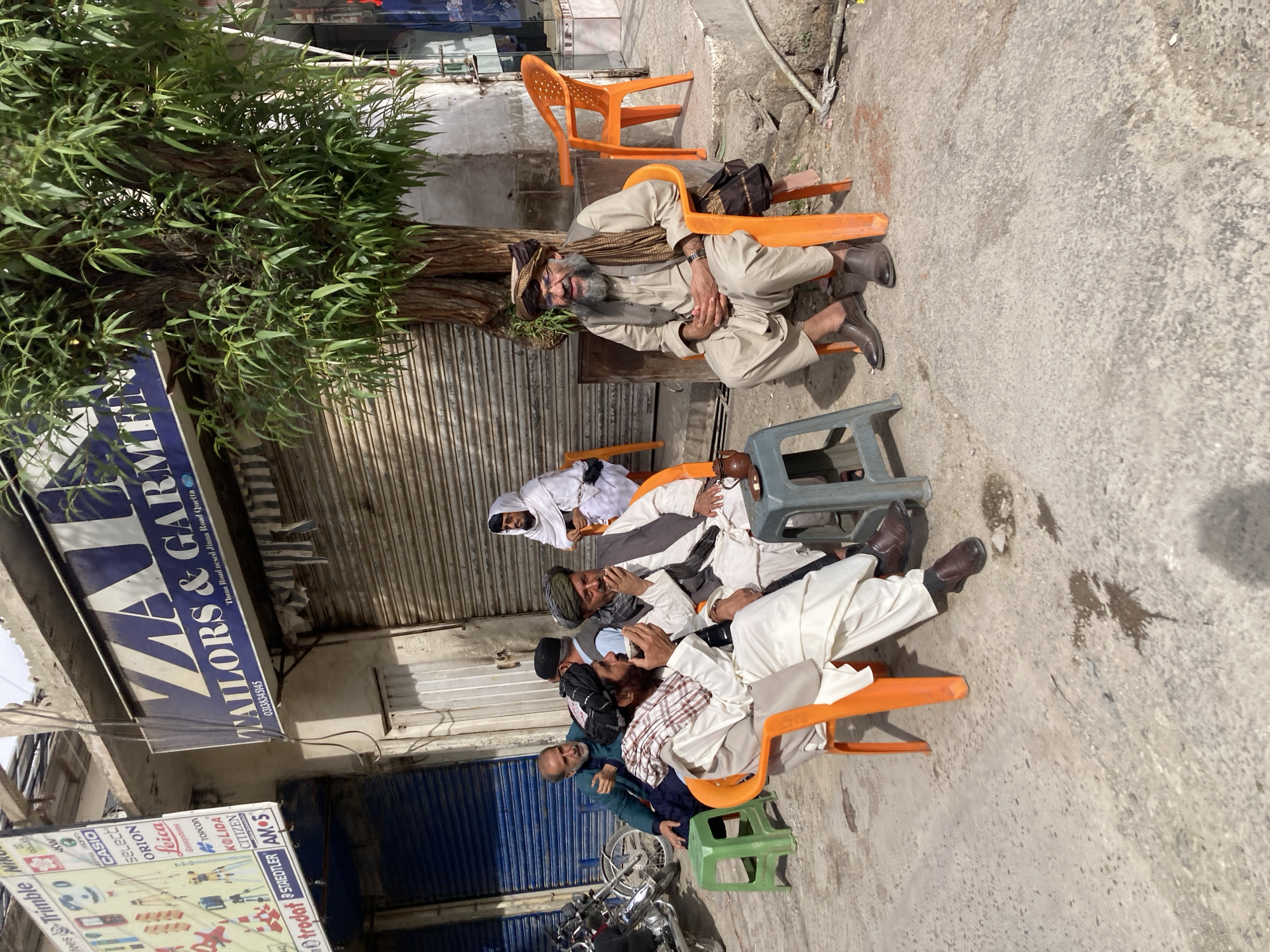 The image shows a group of men sitting on orange and green plastic chairs on a street in Quetta City Tehsil, Pakistan. They are gathered in front of a building with closed metal shutters. A tree stands to the left of the group. A sign above the shutters reads "ZAVLE WAILORS & GARMER," followed by a phone number and location information. In the background, a motorcycle is visible. The men appear to be resting or socializing.
