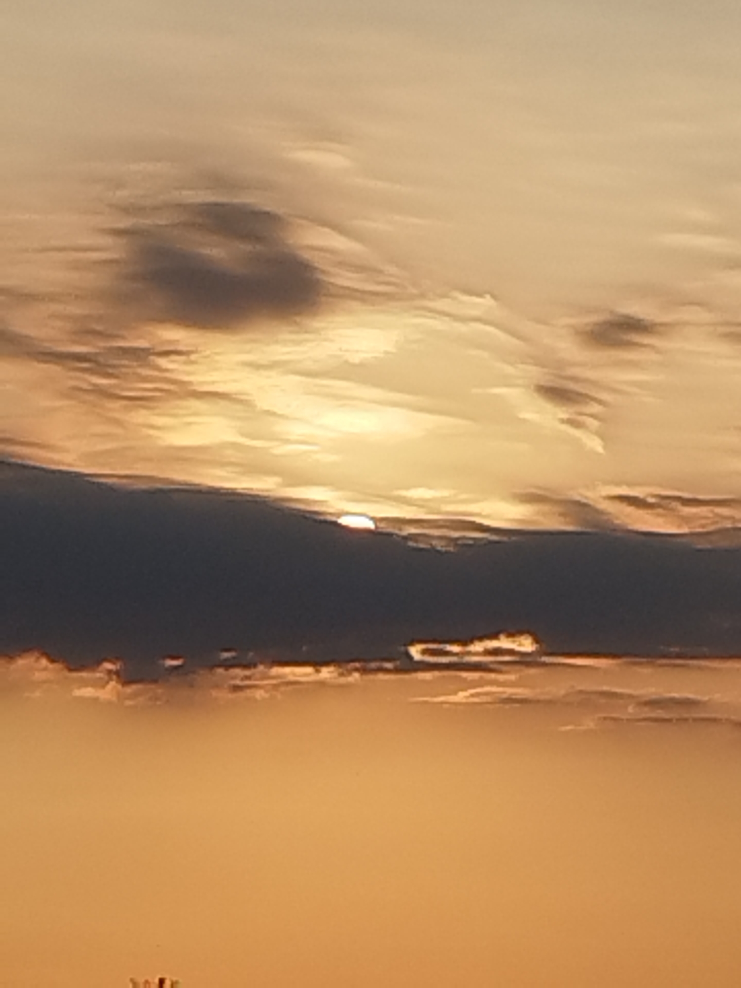 The image captures a sunset over the city of Vidra, Romania. The sky is filled with clouds, ranging in color from golden yellow to dark grey. The sun is partially visible, peeking through a gap in the dark clouds. The lower portion of the image is a soft orange, suggesting the lower atmosphere is illuminated by the setting sun. There are no people or specific objects clearly identifiable in the scene, except for a faint suggestion of structures at the very bottom of the image, which could be buildings. The overall atmosphere is tranquil and serene, typical of a sunset.