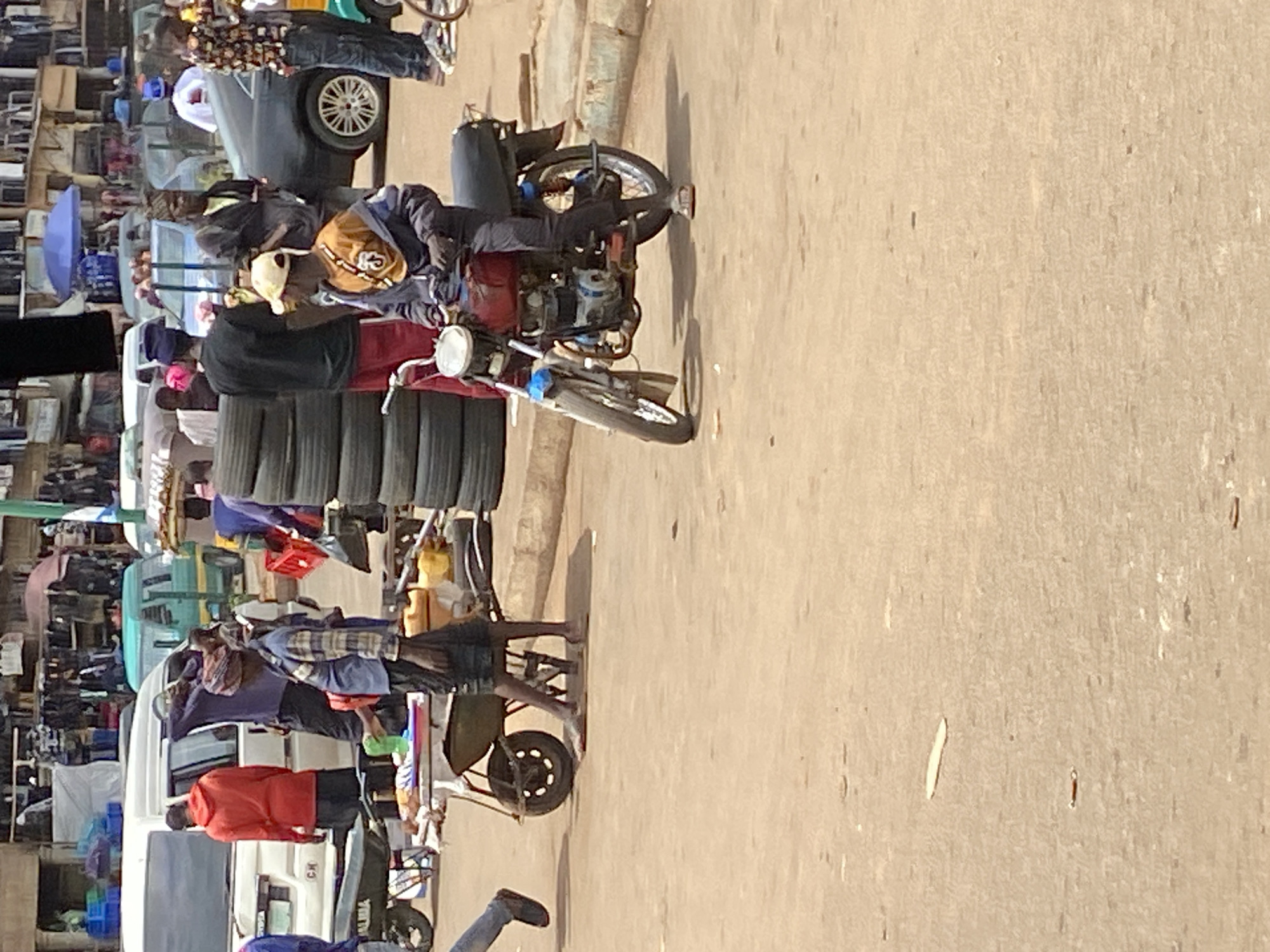 The image depicts a bustling street scene in Bukuru, Nigeria, during what appears to be daytime, indicated by the strong shadows cast on the light-colored, dusty road surface. The weather is sunny and clear.
In the mid-ground, a person wearing a light-colored cap and dark clothing is riding a red and black motorcycle, moving along the road. To their left, another individual is pushing a cart heavily laden with a tall stack of black vehicle tires, also navigating the street.
The left side of the frame reveals a vibrant market or roadside commercial area. Numerous people are visible, either walking, standing, or interacting with goods. Several vehicles are present, including a dark grey car with visible wheel spokes, a white van or light truck, and other partially obscured vehicles. Among the market stalls, a bright blue umbrella stands out. Various other objects like crates, containers, and merchandise contribute to the busy atmosphere.
On the rear of the white vehicle, partial text "CK" is discernible. The overall scene conveys a sense of active commerce and daily life in a lively urban or semi-urban setting.