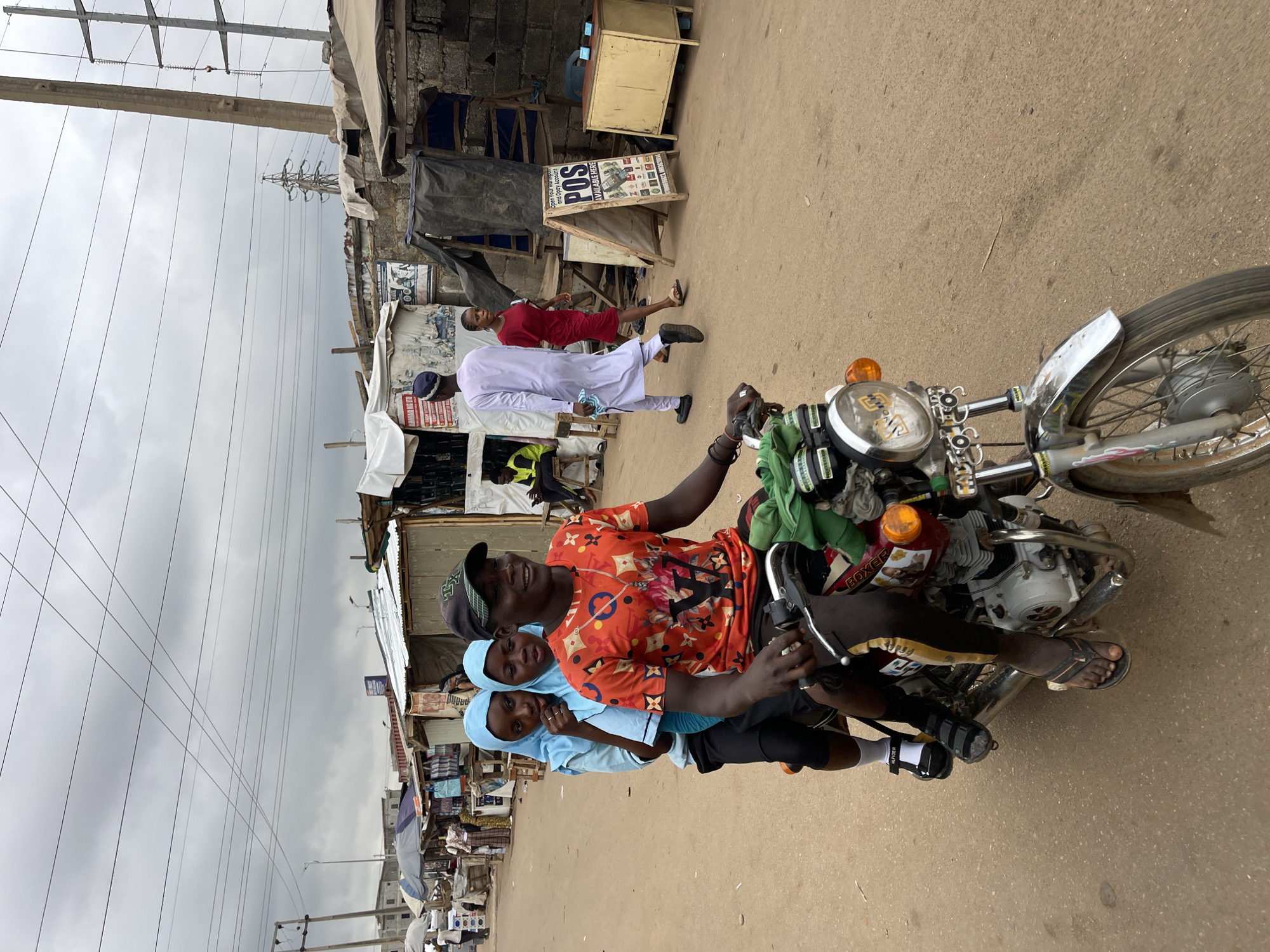 The image shows three people on a motorcycle in Kuchingoro, Nigeria. The motorcycle is traveling on a paved road. The rider is a young man wearing a black baseball cap, an orange shirt with patterns, and black shorts. Two young girls wearing light blue hijabs and shirts are seated behind him. The background includes makeshift structures and shops lining the street. Two individuals are walking on the side of the road in the background. Overhead power lines are visible against a cloudy sky. A sign with "POS" is displayed in one of the buildings.