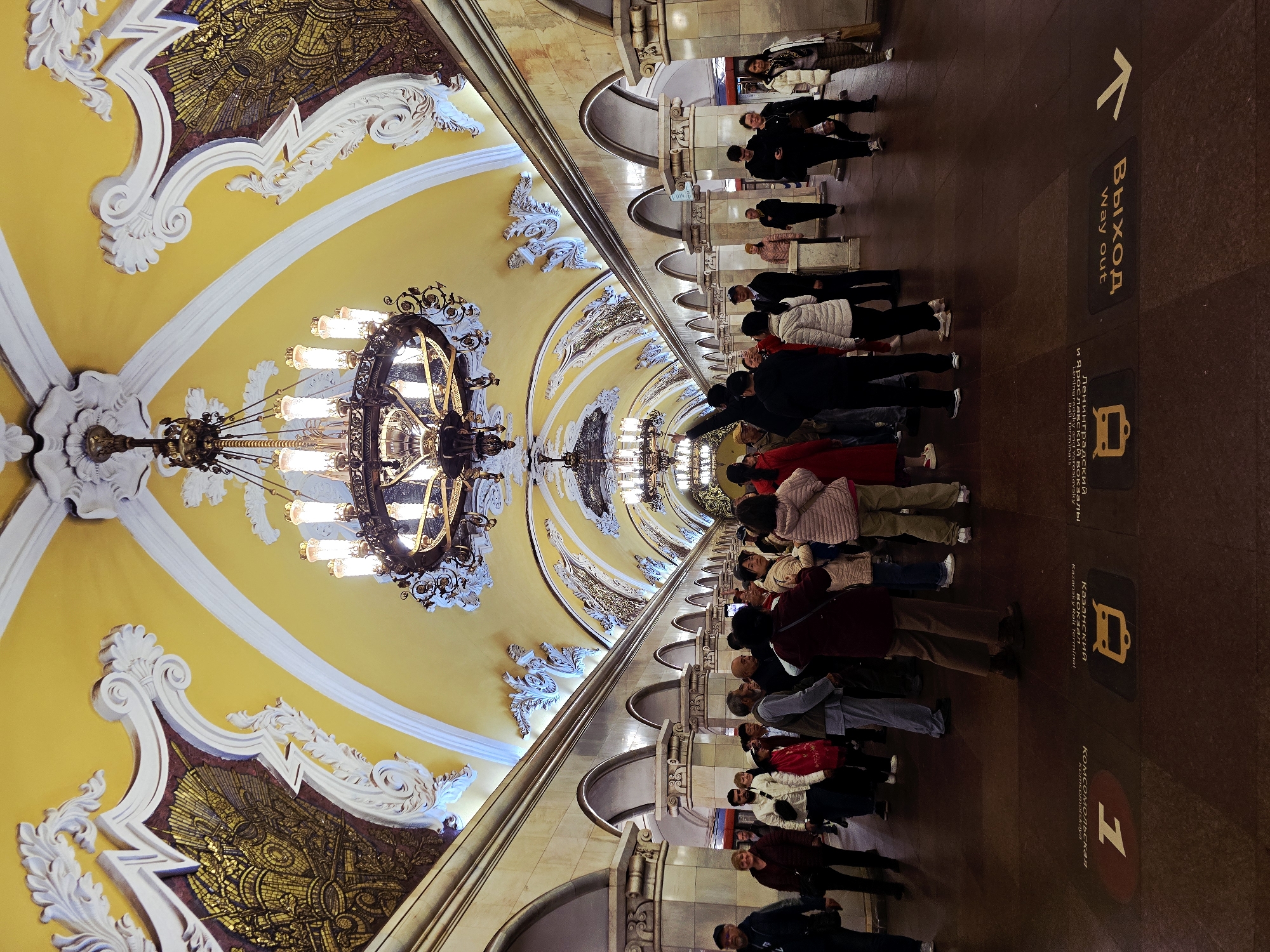 The image shows a subway station in Moscow, Russia. A large group of people are walking to the right along the station platform. Above them is an ornate ceiling with a pale yellow background, white trim, and gold mosaic sections. Several large chandeliers hang from the ceiling. Signage on the floor directs people towards the “Way Out” and towards the "Komsomolskaya" station and Kazansky Railway Terminal.