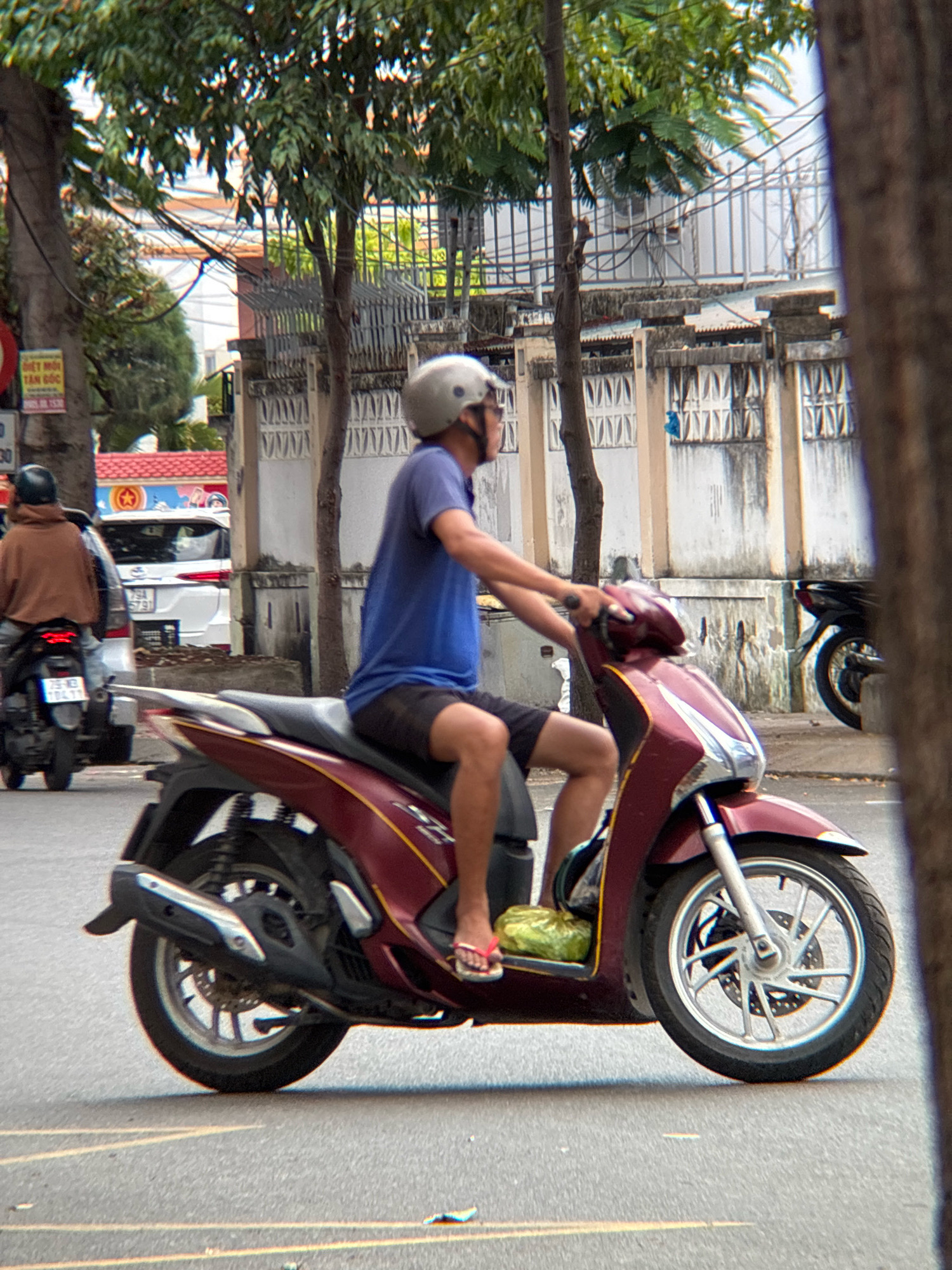 A person wearing a blue t-shirt and dark shorts rides a maroon scooter. The scooter has a silver exhaust pipe and silver rims. The rider is wearing a silver helmet and pink flip-flops. A yellow bag is visible on the floorboard of the scooter. In the background, another person on a scooter wears a brown hooded jacket and a black helmet. A white car is also visible behind them. The scene appears to be a street in Nha Trang, Vietnam, with trees, buildings, and a wire fence in the background. The road is paved and has yellow lane markings.