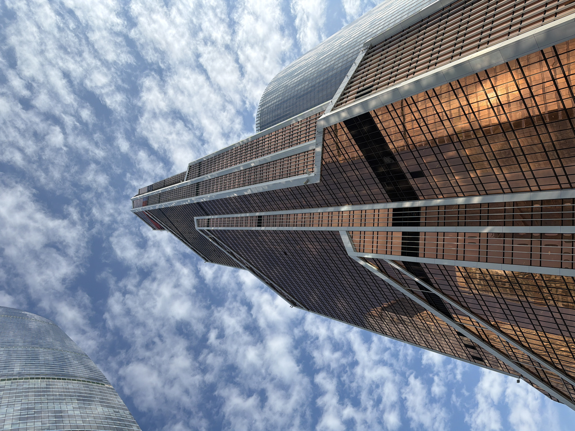 The image captures a low-angle view of the architectural elements of buildings in Moscow, Russia, emphasizing the interplay of modern design with the natural sky. The focus is on the upward perspective, highlighting the height and scale of these structures against a backdrop of a blue sky interspersed with white clouds. The buildings feature reflective facades, showcasing a grid pattern of windows. The composition frames the sky with modern architecture.