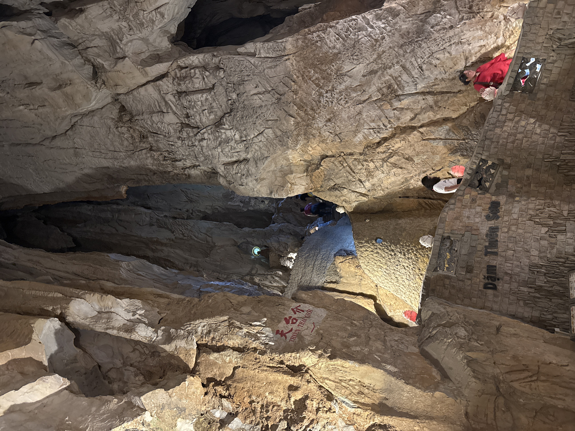 This image depicts a section of a large, naturally formed limestone cave, identified as part of the Marble Mountains (Ngũ Hành Sơn) in Đà Nẵng, Vietnam. The scene is illuminated by artificial lighting, revealing rugged, textured rock formations that constitute towering walls and a high ceiling. Man-made structures are integrated into the cave environment, featuring brick-patterned walls and stone staircases.
On a prominent rock face to the left of the center, a sign is painted in red Vietnamese and Chinese characters, reading "天台界" (Thiên Thai Giới), which translates to "Thiên Thai World," accompanied by a red arrow pointing upwards and to the right. A lower, curved brick wall features the partially visible text "DẠM THIÊN SƠN" (Dam Thien Son) in dark letters.
Approximately five individuals, appearing to be tourists, are present. In the lower right foreground, a person wearing a red shirt or jacket stands on a curved brick ledge, holding a white, lotus-shaped object. To their left, on a parallel but slightly higher brick ledge, another person in a white t-shirt and dark pants stands beside a red lotus-shaped lantern. Further into the cave, a stone staircase ascends upwards and to the right, where three additional individuals are visible in various stages of ascent. One of these individuals wears a light-colored backpack, and another wears dark clothing with red pants. High up within the cave, two green and one blue circular accent lights are visible against the dark rock. The artificial illumination creates dramatic shadows and highlights the geological features and the integrated pathways.