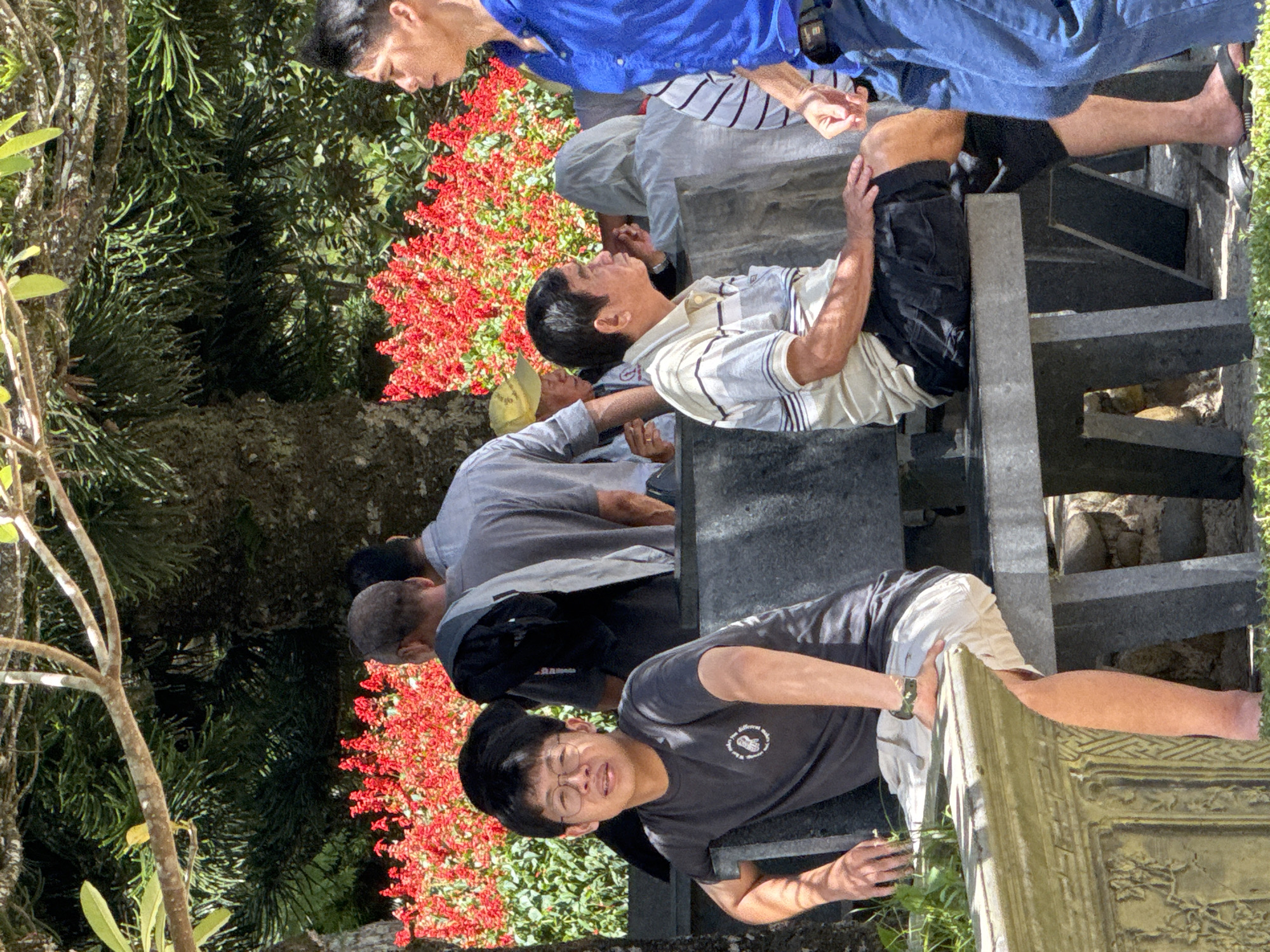 A group of men are gathered outdoors, near a stone structure that appears to be part of a tomb or monument. The setting is lush with greenery and vibrant red flowers. The time of day appears to be sunny, suggesting daytime. The men are dressed casually, some in t-shirts and shorts, while others wear collared shirts. They are positioned around the stone structure, with some sitting and others standing. One man in the foreground is looking towards the camera, his mouth slightly open, suggesting he might be speaking or reacting to something. Another man, wearing a blue shirt, is partially visible at the top of the image. The overall mood seems relaxed, possibly a social gathering or a moment of reflection. There is some text visible on the t-shirt of the man in the foreground, but it is too small and pixelated to be legible. The scene evokes a sense of community and shared experience in a natural, contemplative environment, characteristic of locations like Da Lat, Vietnam, known for its gardens and serene atmosphere.