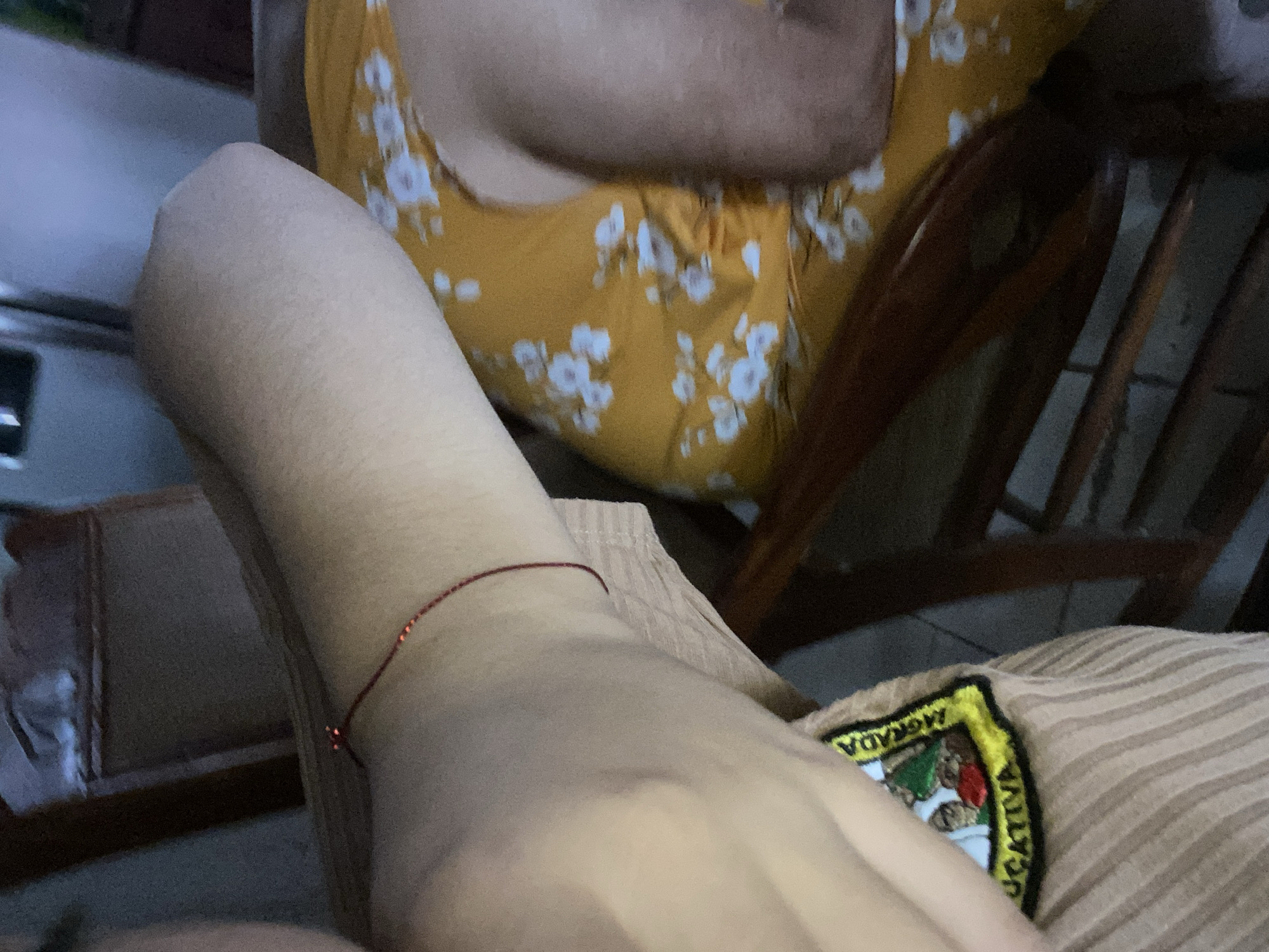 The image shows a person's arm resting on their lap. A red string bracelet is visible on the wrist. The person is wearing a beige striped garment with a patch that reads "MORADA EDUCATIVA." In the background, another person is seated, wearing a yellow floral dress, in a wooden chair. The environment is indoors, possibly a home. There is no information related to Cotoperíz, Venezuela.