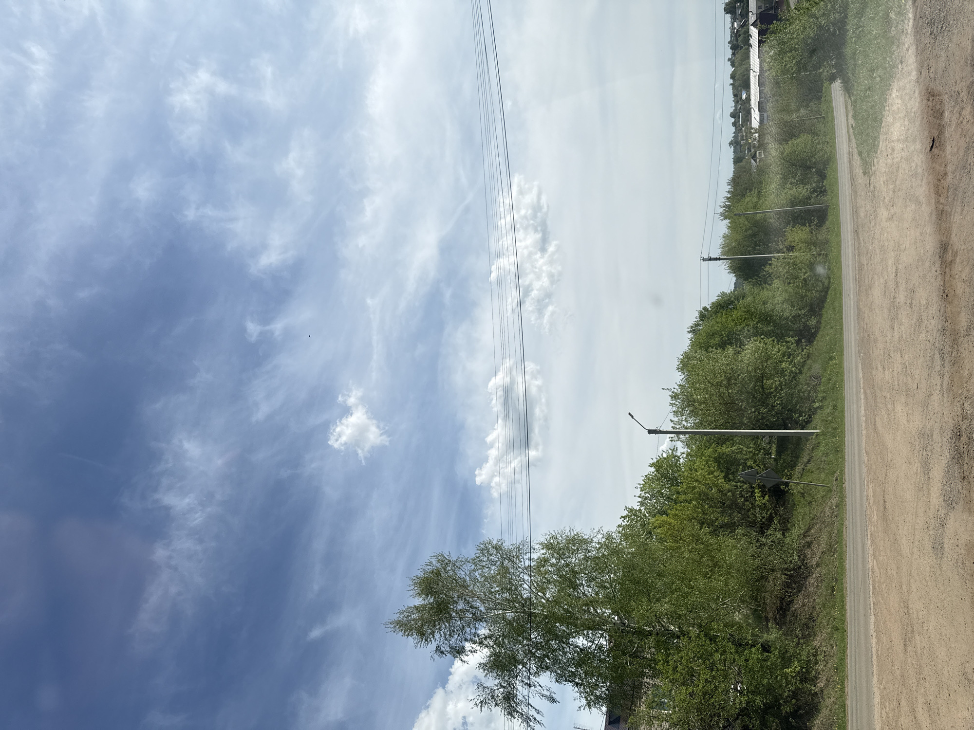 The image shows a rural scene likely captured in or near Фоки, Russia. A dirt road runs along the right side of the frame, bordered by a grassy area and trees. Utility poles line the roadside, with power lines stretching across the sky. In the distance, buildings and more vegetation are visible. The sky takes up a significant portion of the image, displaying a mix of blue and white clouds. A birch tree is visible in the foreground.