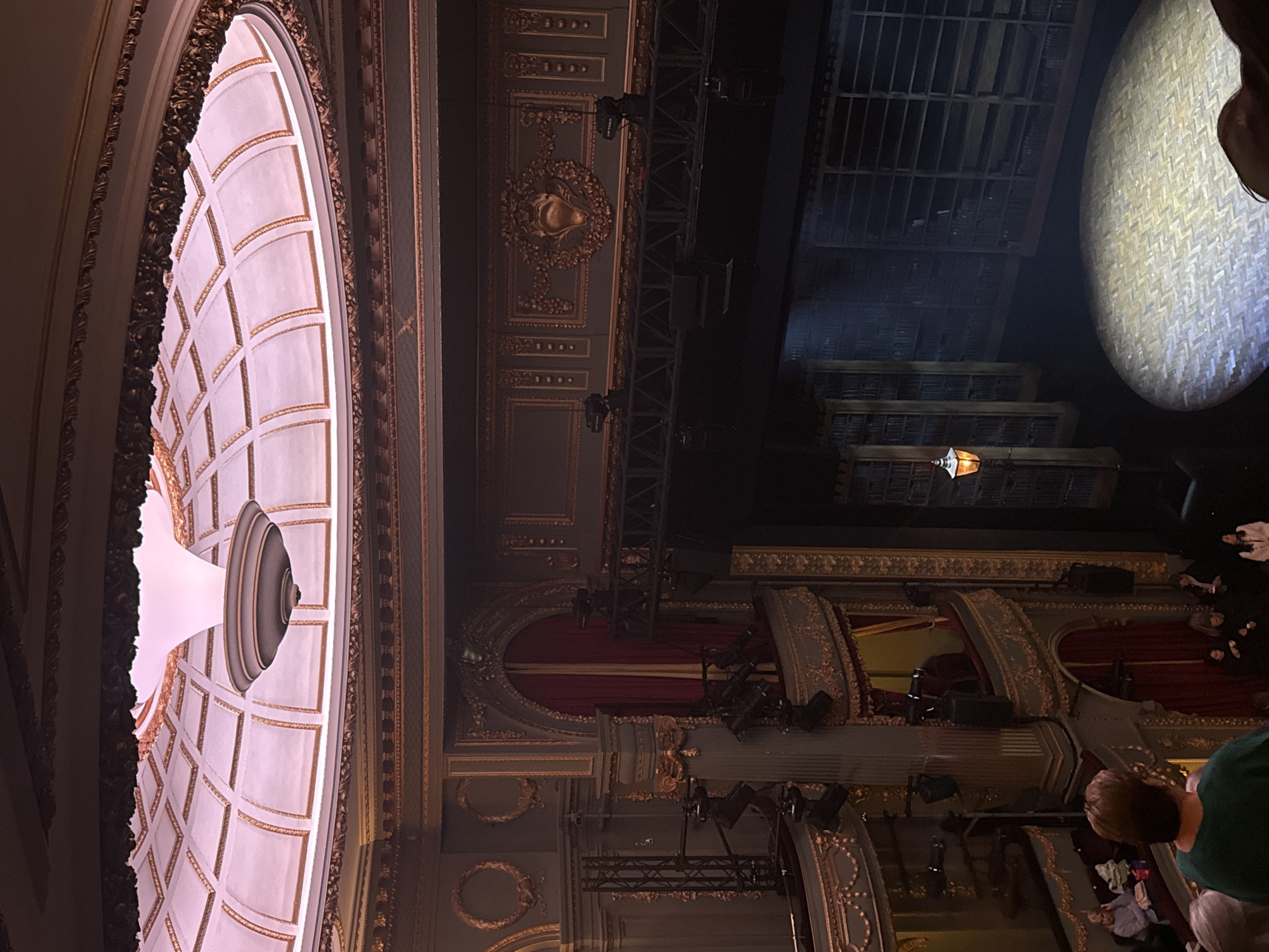 The image shows the interior of a theater in London, United Kingdom. A large ornate ceiling with detailed architectural features and decorative molding is visible. Stage lighting and rigging are suspended above the stage area, which features red curtains and decorative elements. A section of tiered seating is populated with audience members.