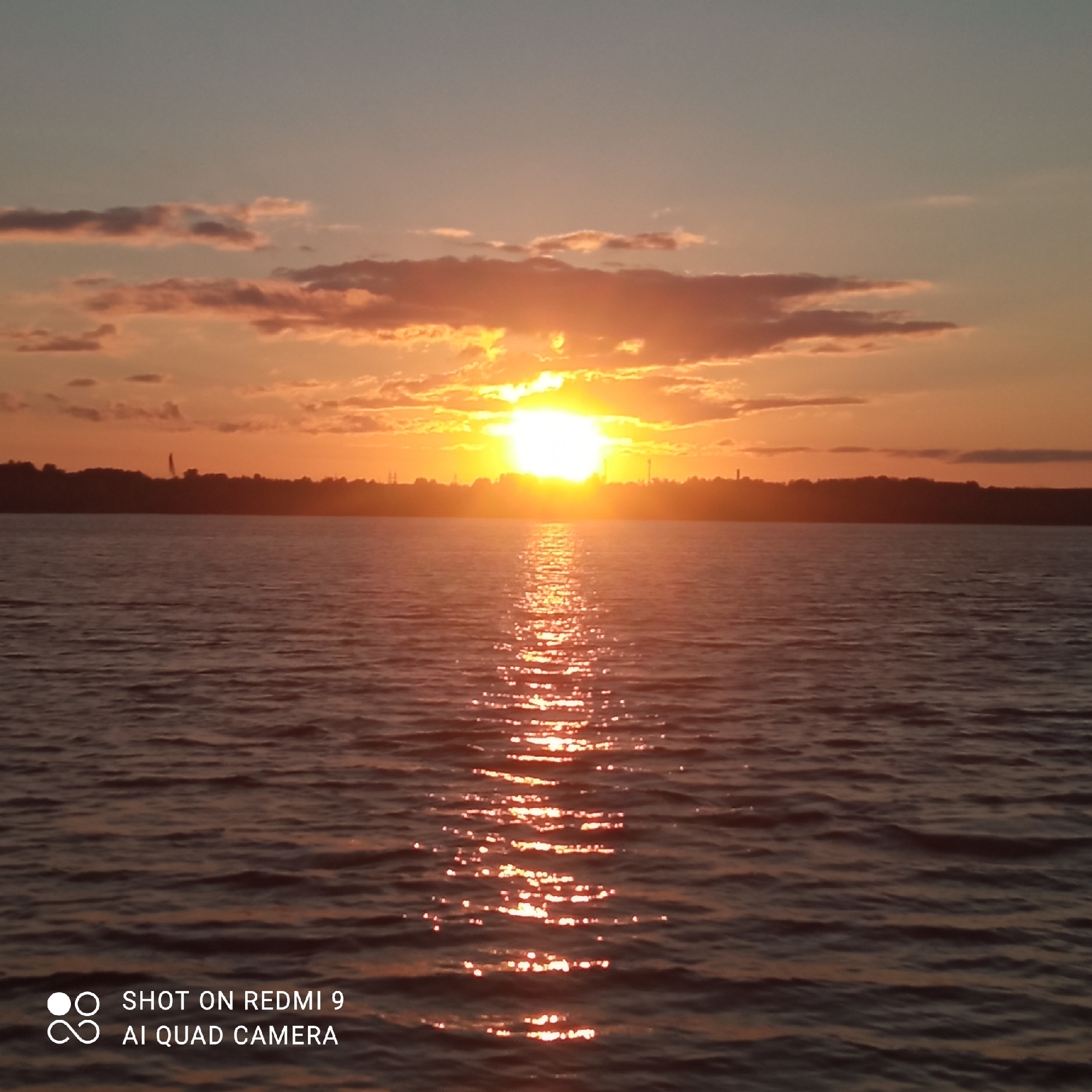 This image depicts a sunset over a body of water, likely the Kama River, near the city of Chaikovsky, Russia. The sky is filled with warm hues of orange and yellow, with scattered clouds tinged pink and purple. The sun, a bright orb, is partially obscured by clouds on the horizon. A reflection of the sun shimmers on the rippling surface of the water. In the distance, the silhouette of the city's shoreline is visible, featuring trees and some structures, including what appears to be a tall tower or industrial chimney. The lower portion of the image contains a watermark indicating "SHOT ON REDMI 9 AI QUAD CAMERA". There are no people or specific actions depicted.
