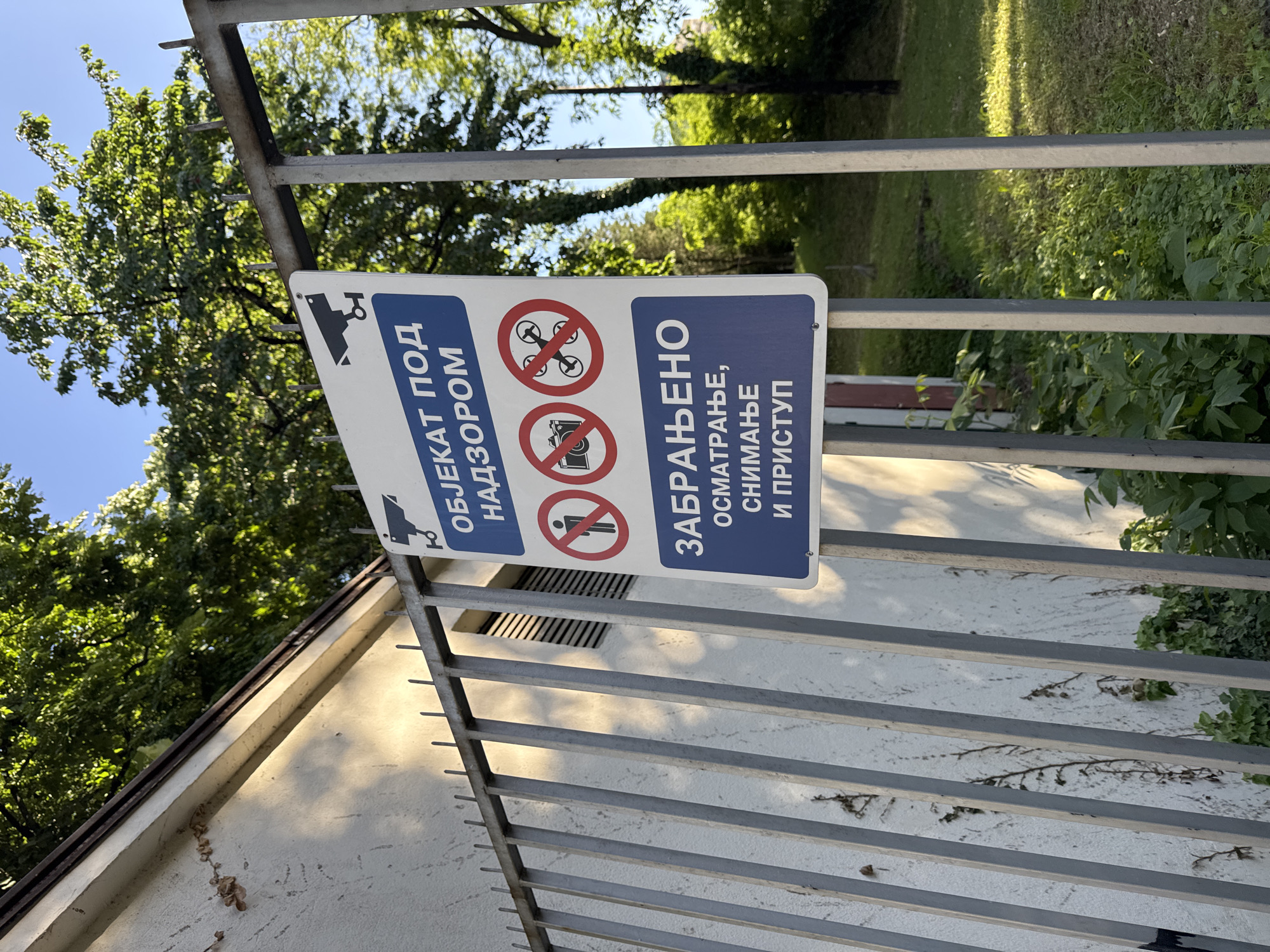 A sign is attached to a metal gate in Vracar Urban Municipality, Serbia. The sign is blue and white with Serbian text and symbols. The text reads "ОБЈЕКАТ ПОД НАДЗОРОМ" (Object Under Supervision), "ЗАБРАЊЕНО ОСМАТРАЊЕ, СНИМАЊЕ И ПРИСТУП" (Forbidden Observation, Filming and Access). The symbols include a surveillance camera, a drone with a red circle and line through it, a camera with a red circle and line through it, and a person with a red circle and line through it. The gate is grey and has horizontal bars. Behind the gate are trees and a building. The sky is blue.